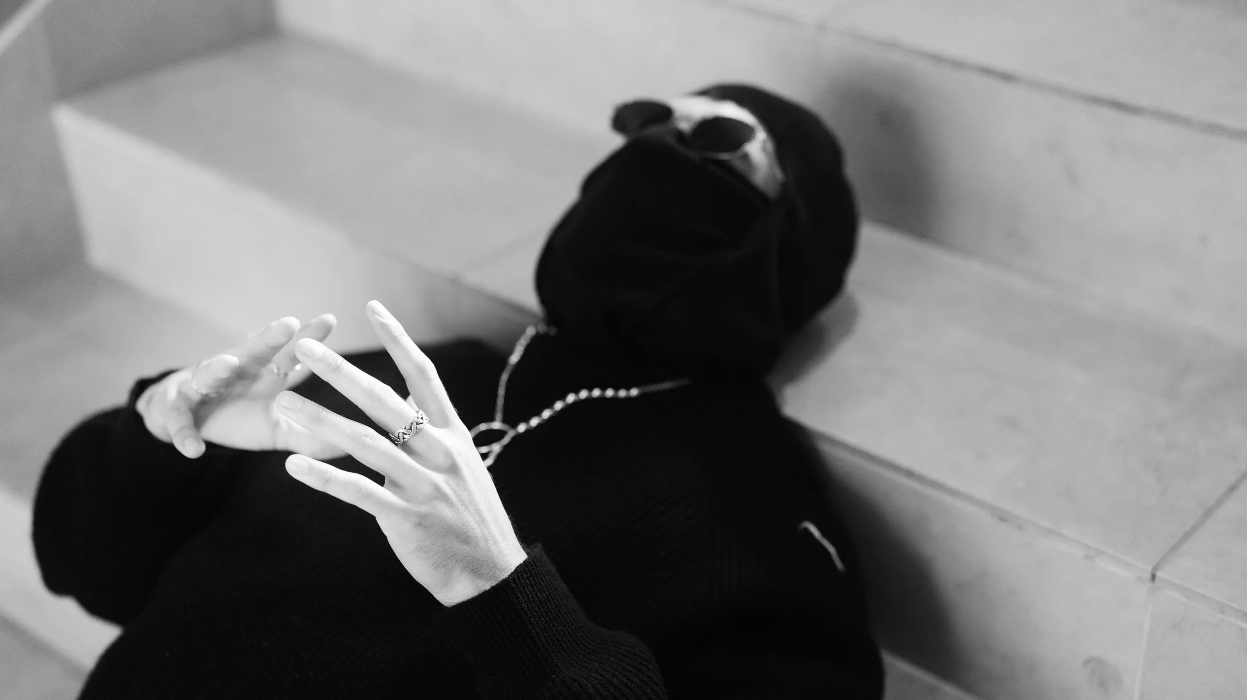 Black and white photo of a person's hand with multiple rings and a chain necklace, resting on their lap, with a black bag in the background.