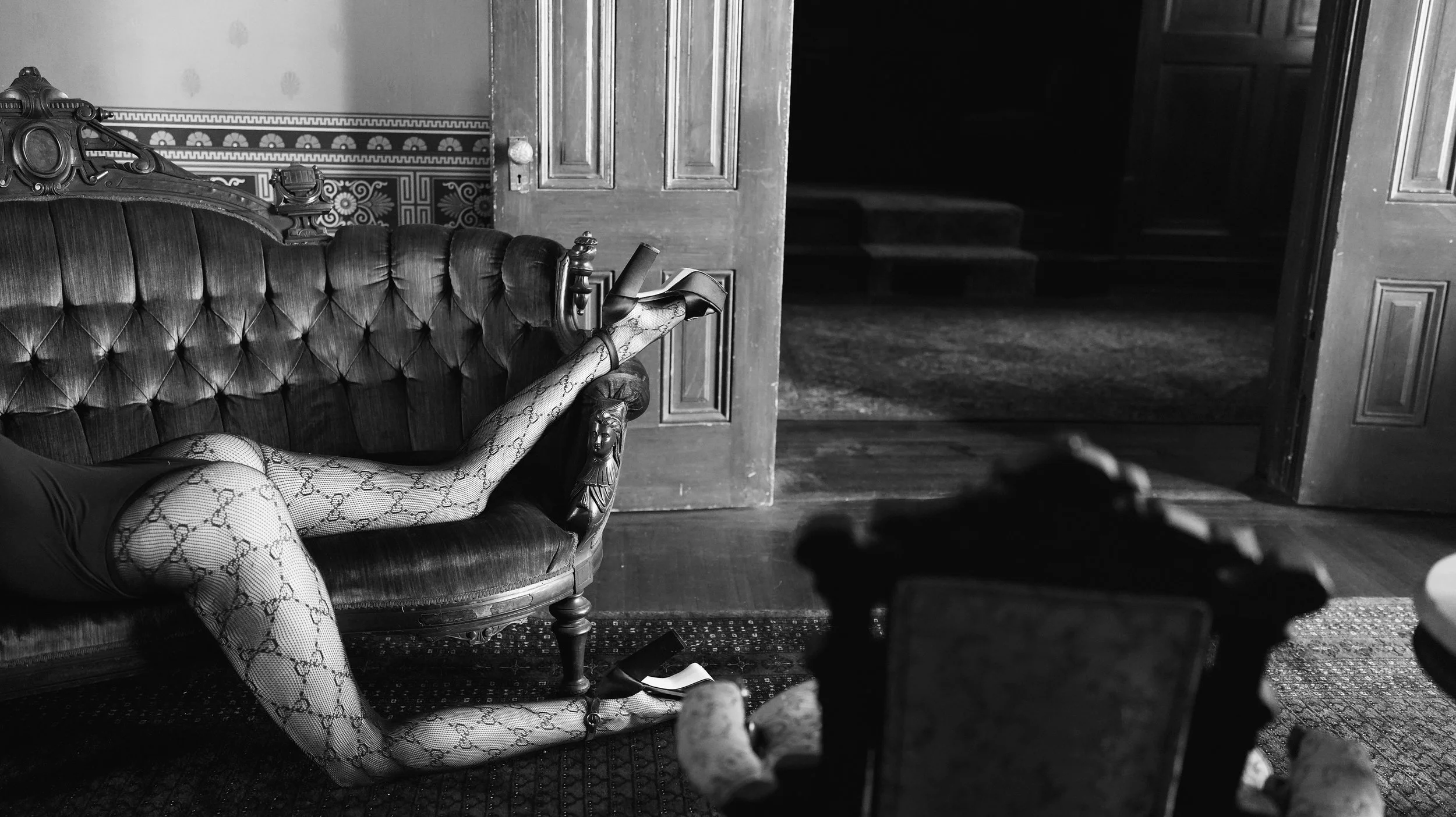 A woman wearing patterned tights and high heels lounges on a vintage sofa in an old, ornate room with patterned wallpaper and wooden doors.