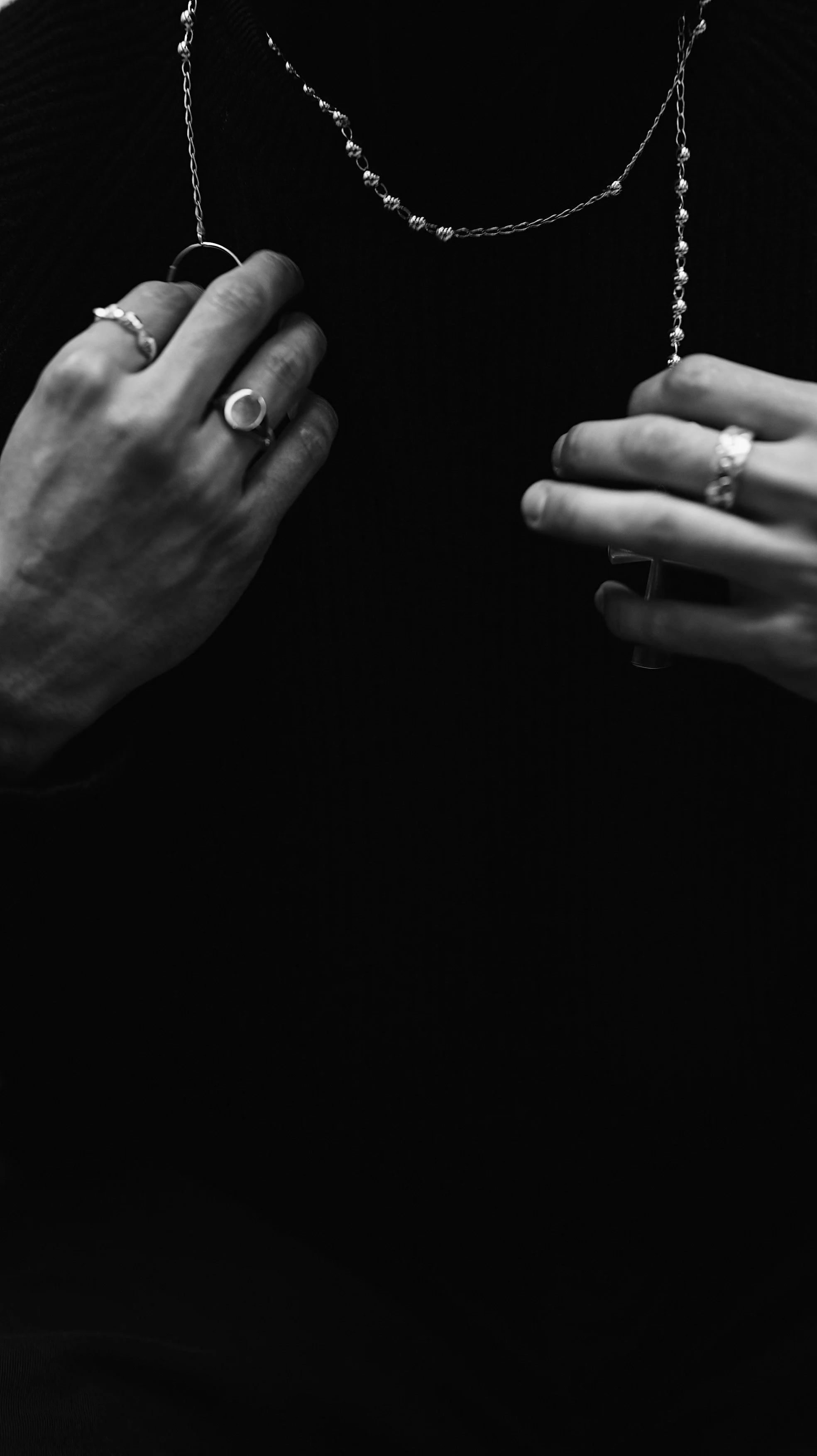 Close-up of a person wearing rings and necklaces while holding two jewelry pieces. The photo is in black and white.