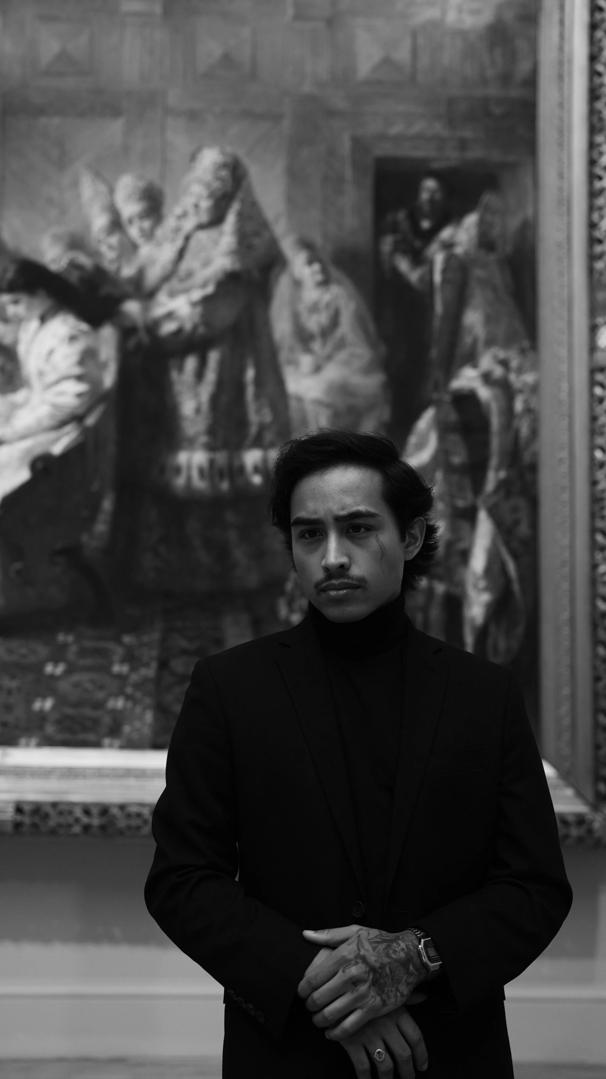 A young man with dark hair and tattoos on his hands standing in front of a large black-and-white painting at an art gallery. He is wearing a black turtleneck and blazer, with his arms crossed.