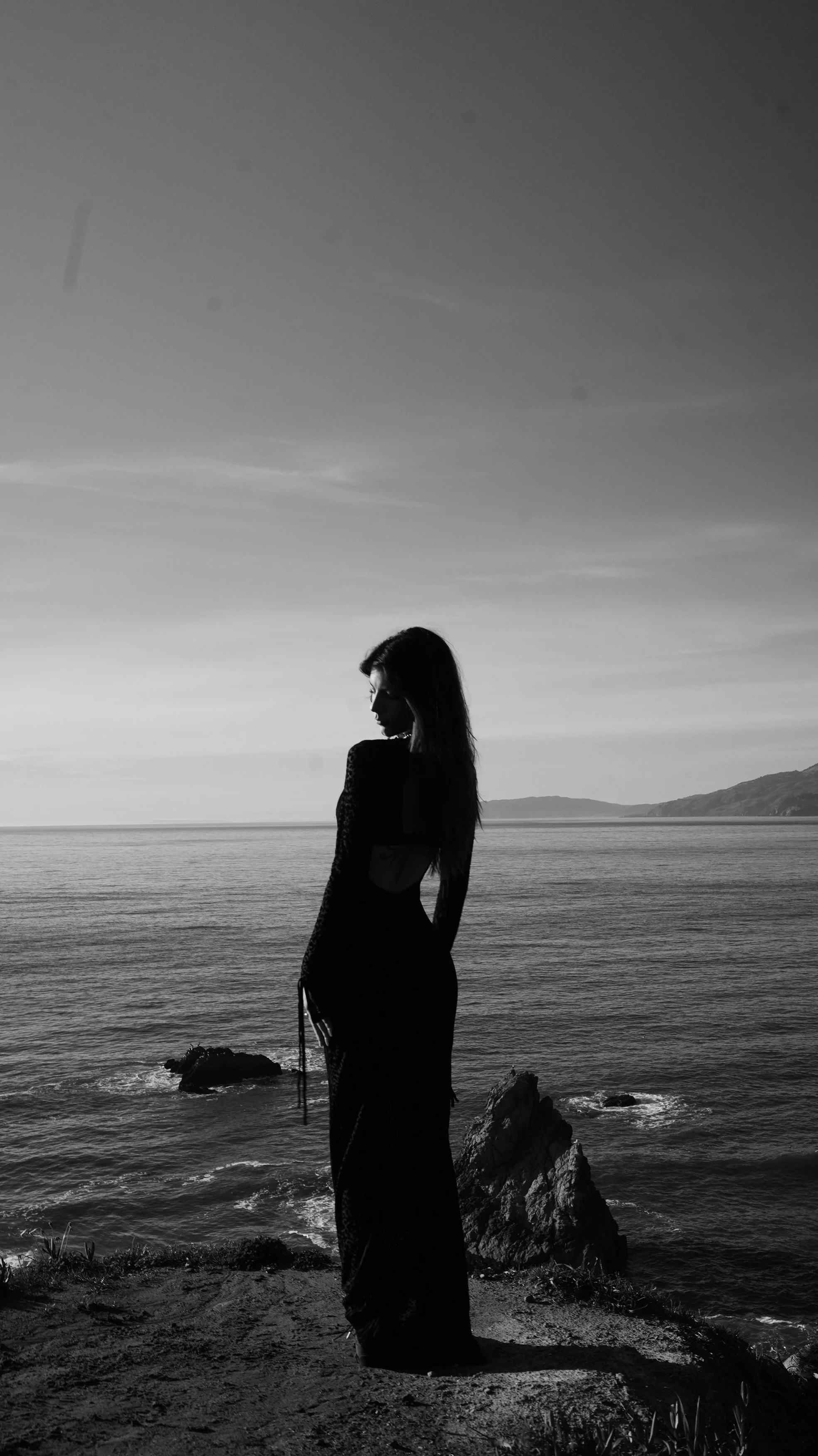 A black and white photograph of a woman standing on a rocky shoreline by the ocean, with a distant landmass on the horizon and the sky above.