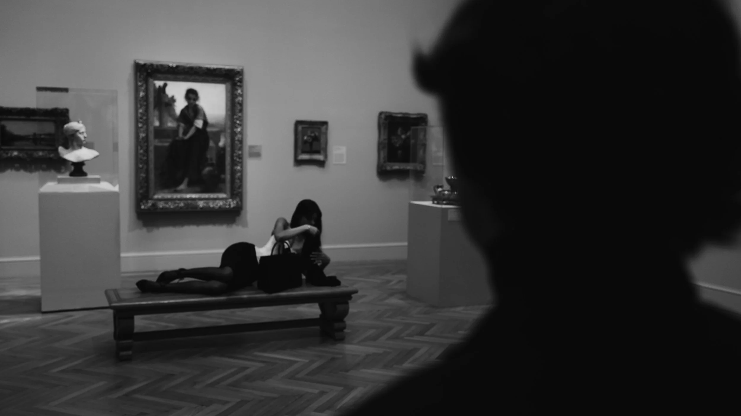 Black and white photo of two young girls lying on the floor in an art gallery, with paintings and sculptures displayed on the walls and pedestals. One girl is on her side, leaning on her arm, while the other is partly obscured. A man's silhouette is 