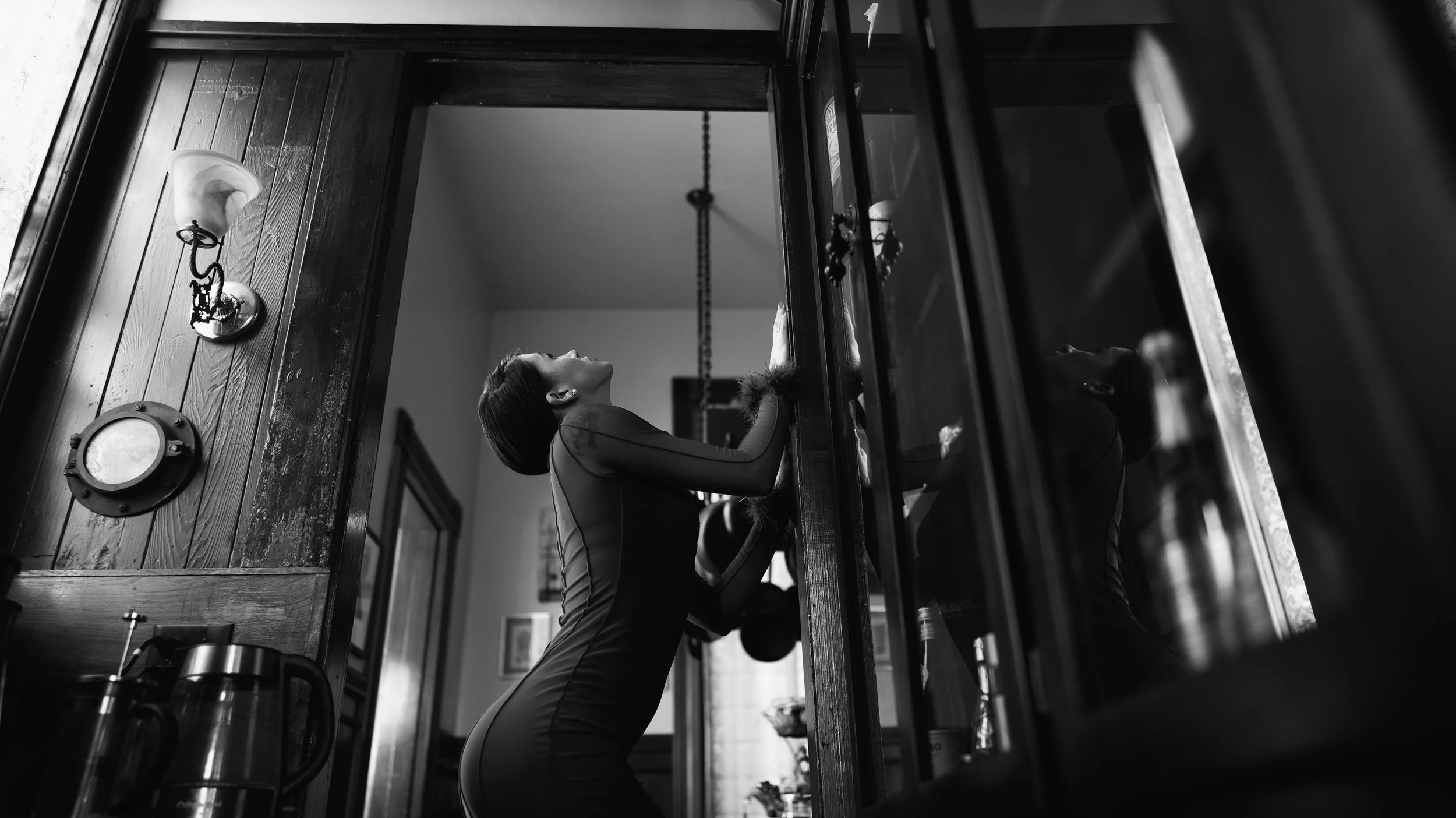 A woman is stretching her arms above her head while leaning on a wooden table in a room with wooden walls, with a mirror and a wall sconce visible in the background.