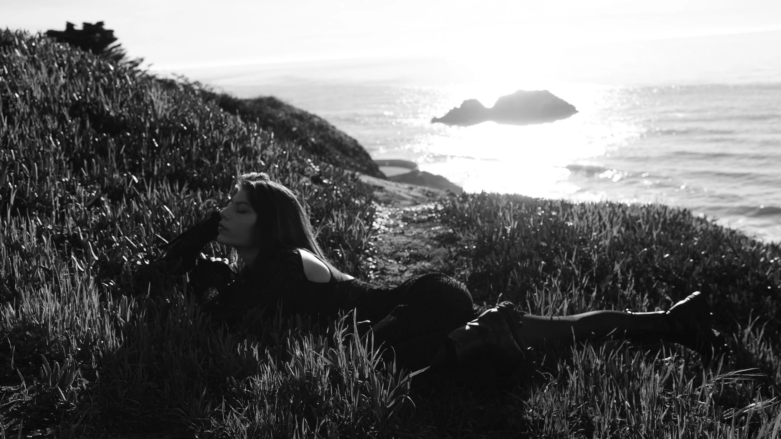 A woman reclining on grass near the coast, with ocean and rocky islands in the background, black and white photo.