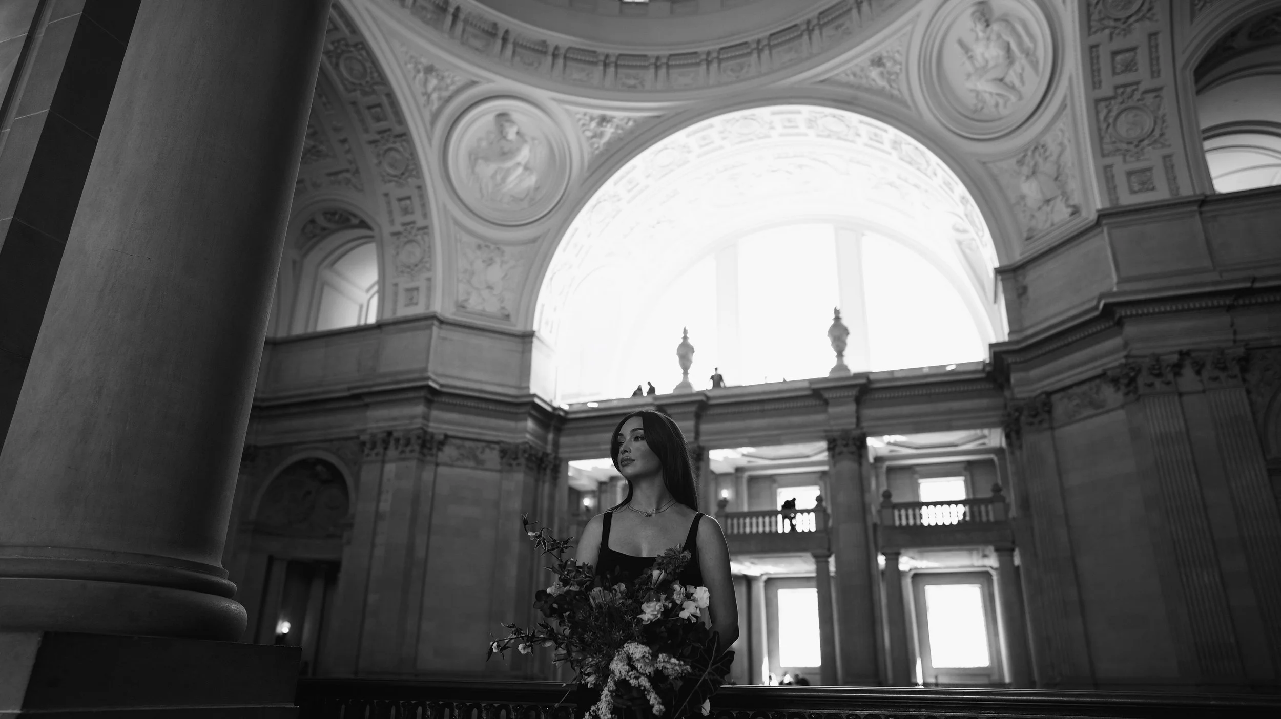 A woman in a black dress holding a large bouquet of flowers inside a grand, historic building with ornate ceiling art and large windows.