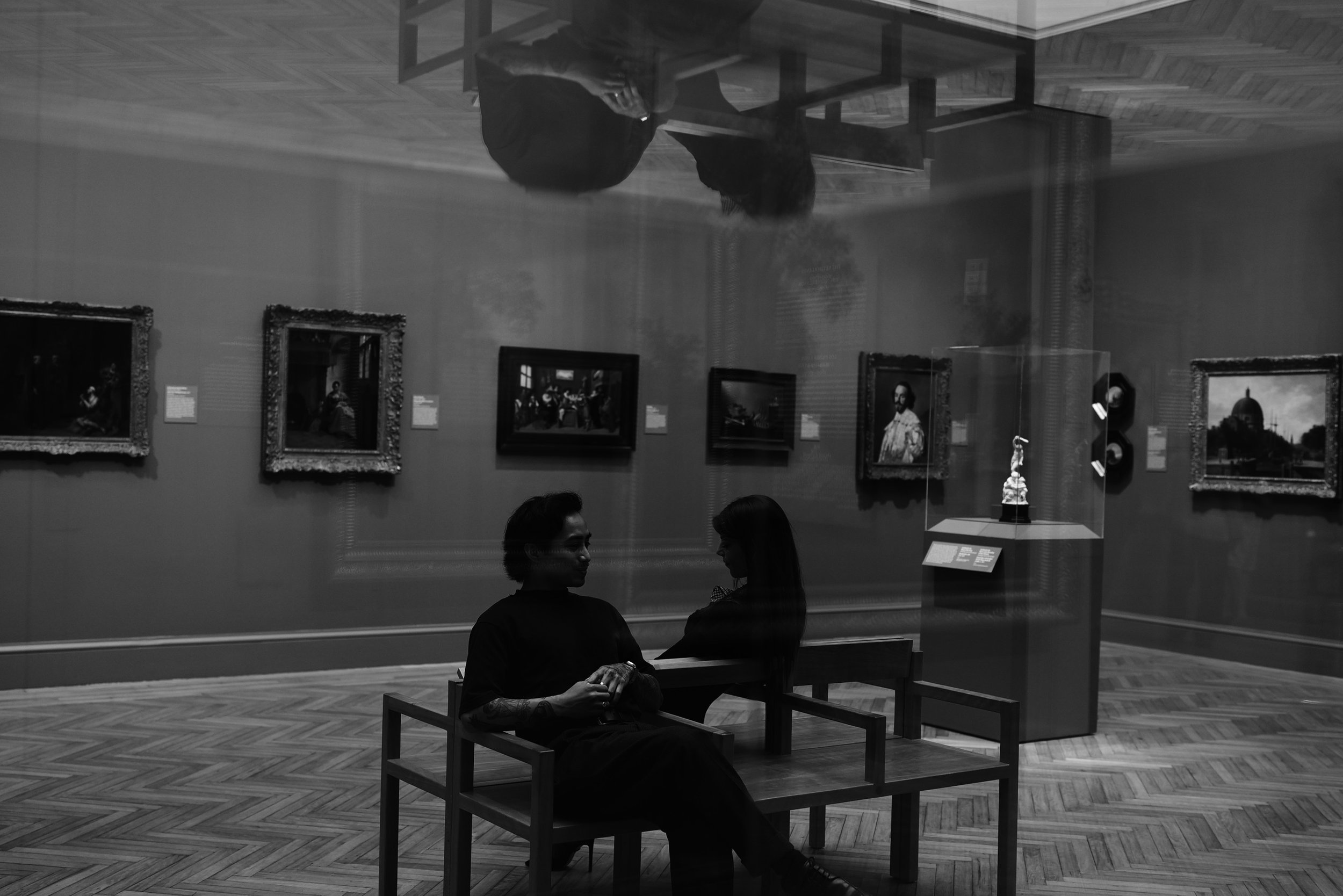 Black and white photograph of two people sitting at a table inside an art museum, viewed through a glass wall that shows framed artwork on the museum's gallery wall behind them.