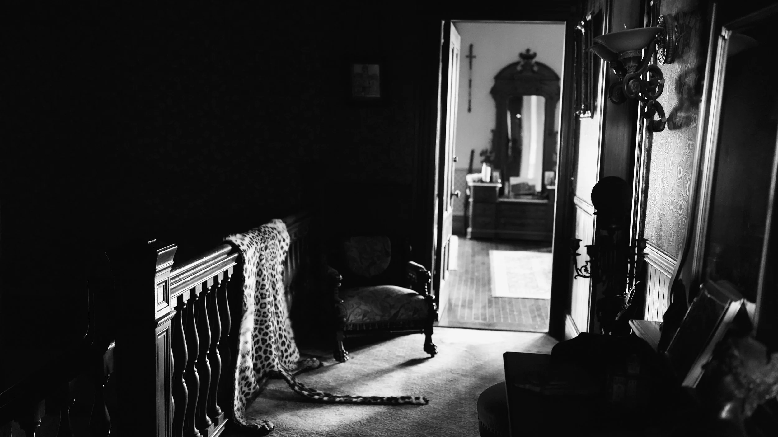 A black and white photo of a hallway in a house. The hallway has wooden furniture, including a bench with a leopard print cloth and a floral chair. An open door reveals a room with a wooden cabinet, a cross on the wall, and framed pictures. Sunlight 