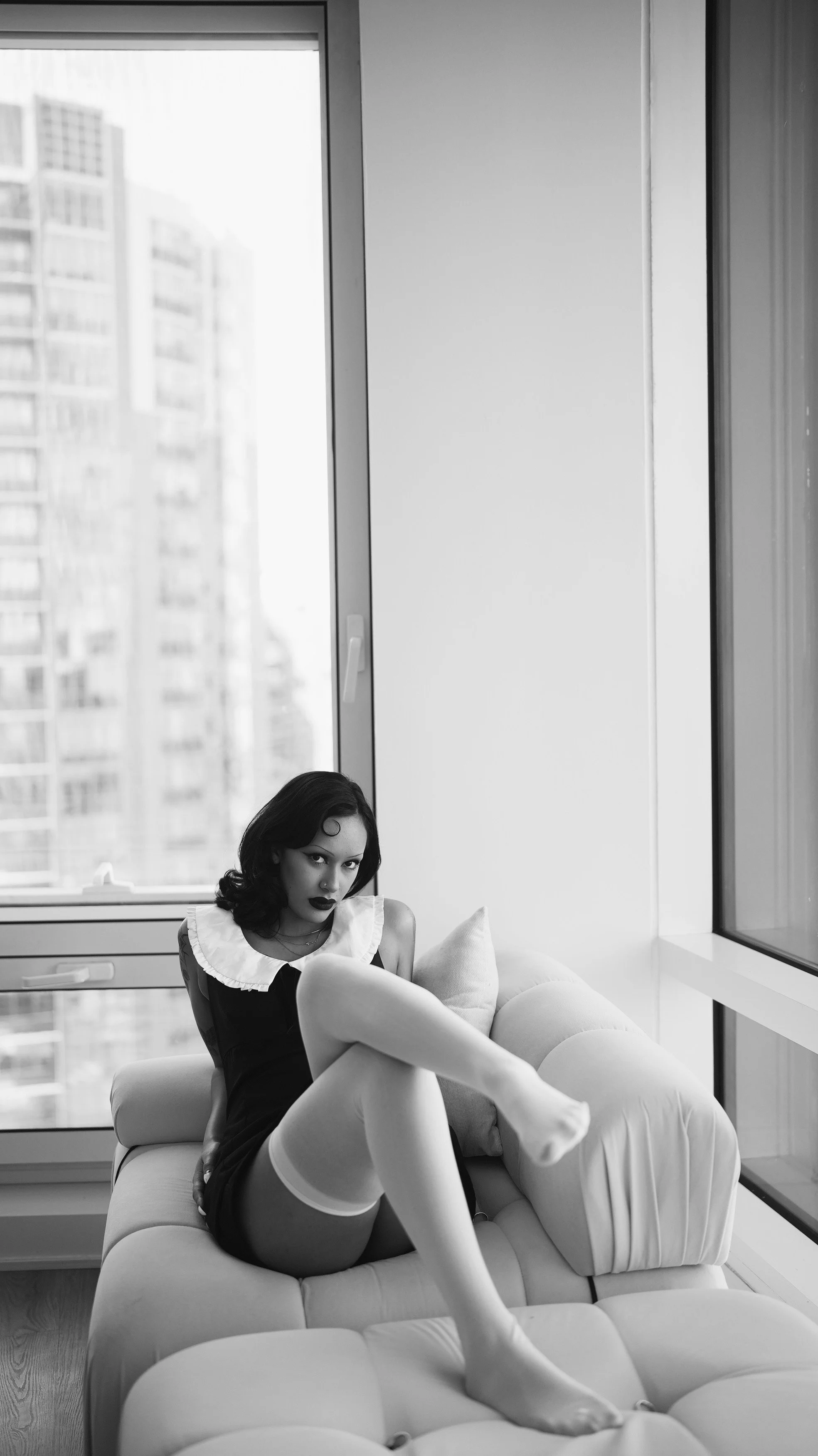 A woman with dark hair, wearing a black dress and white socks, is reclining on a white sofa in front of large windows in a high-rise building. The background shows a cityscape with tall buildings.