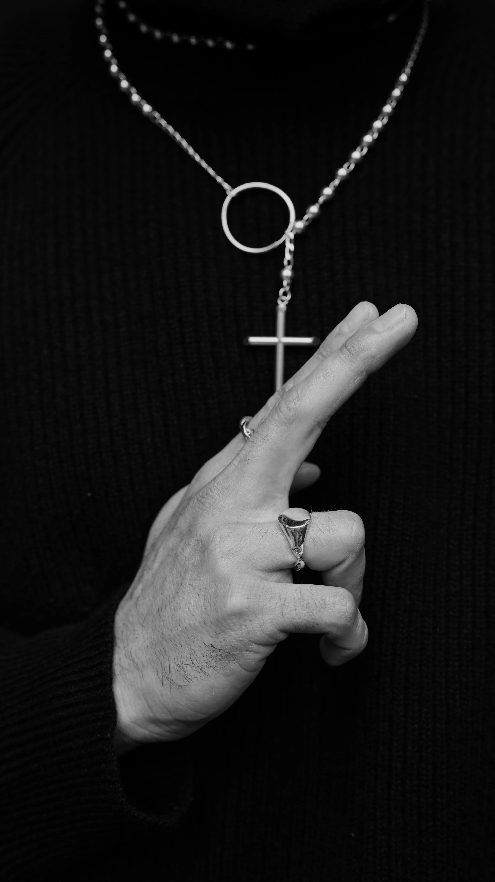 Close-up of a person's hand making a peace sign, wearing rings, with a black shirt and necklaces, including one with a cross and a circle.