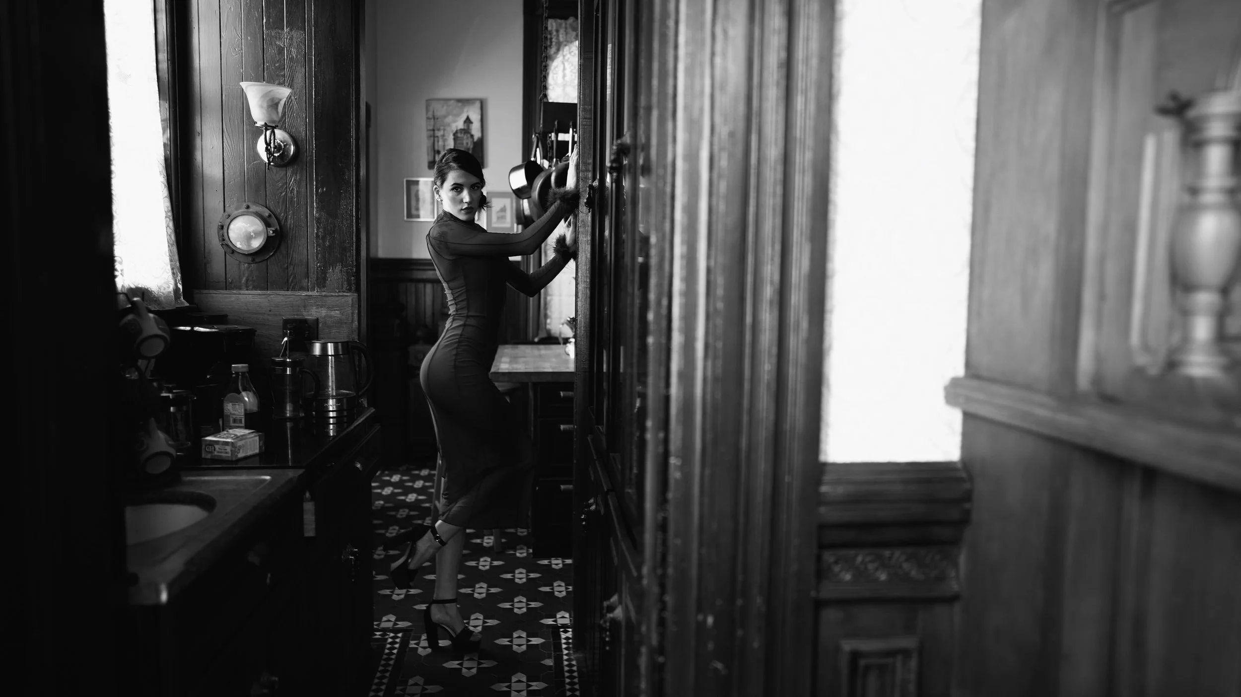 A woman in a fitted dress and heels standing in a vintage kitchen, leaning against a cabinet with her hand on the door, looking towards the camera, with wooden paneling and decorative lighting on the wall.