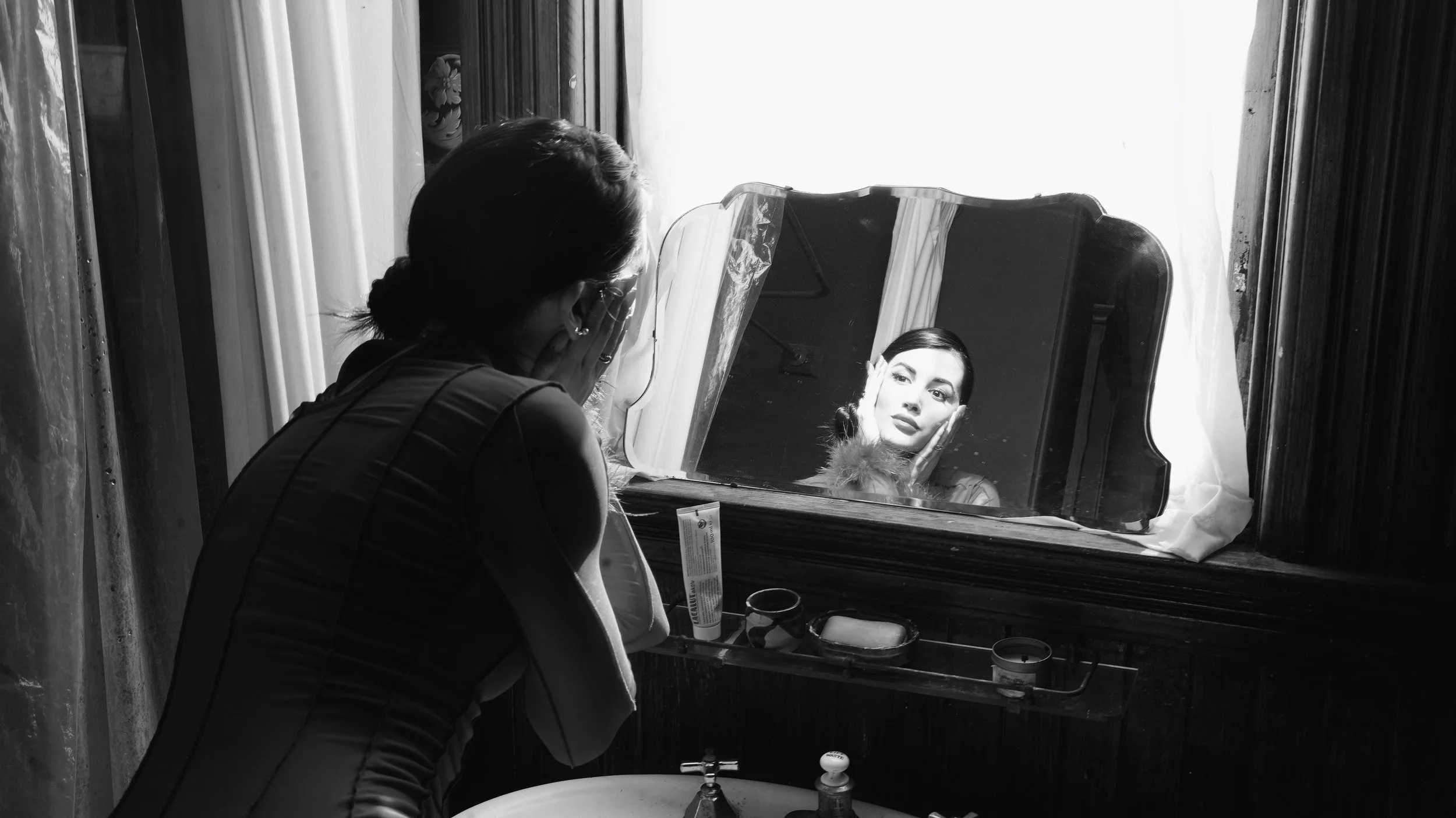 A woman looking at her reflection in a vintage tabletop mirror, sitting at a dressing table in a room with natural light, functioning as her vanity mirror.
