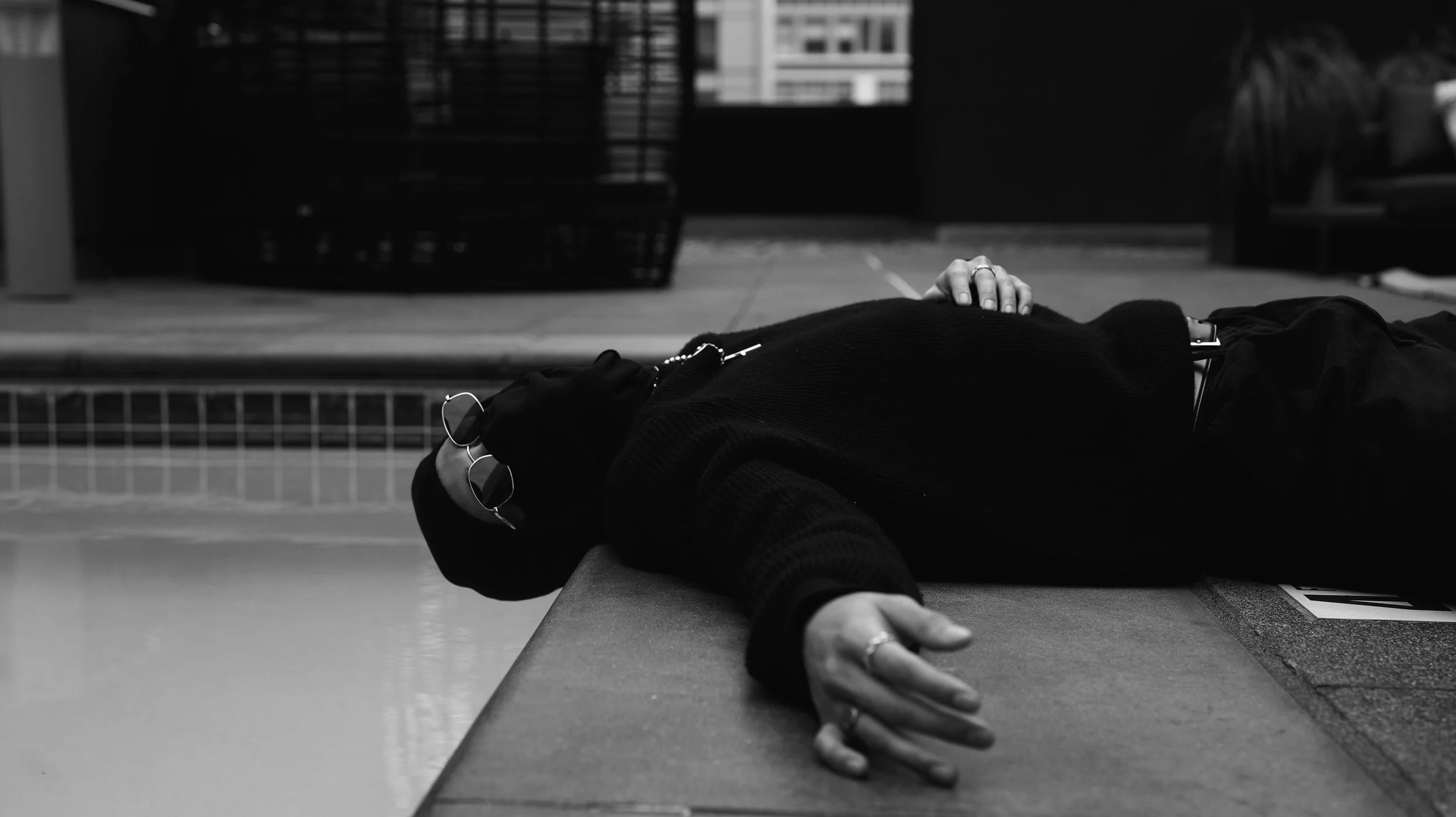 A person lying face down on a table or flat surface with their arm extended forward, wearing glasses, a ring, and dark clothing. The background shows an indoor space with a cage and window.