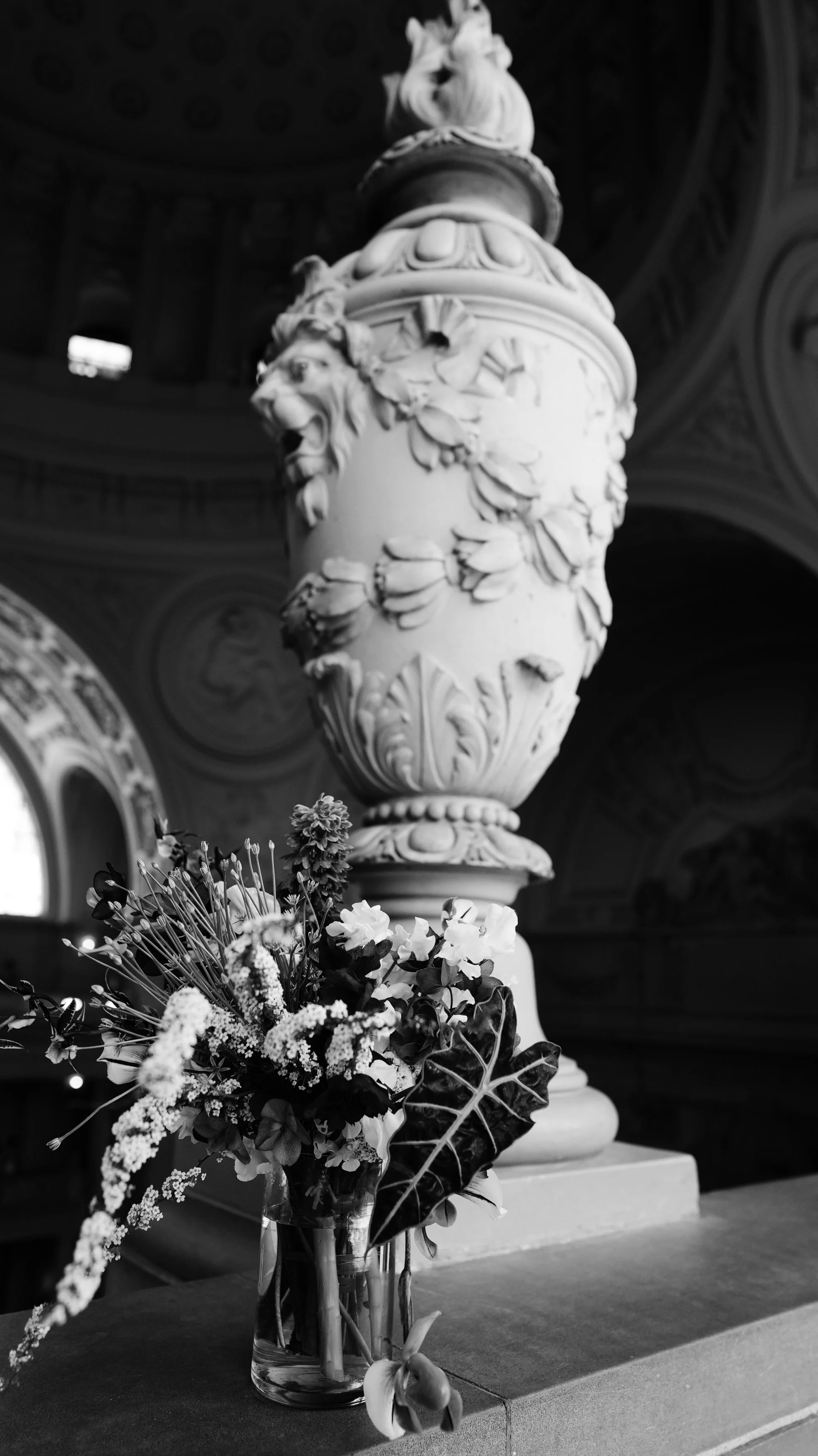 A decorative urn with intricate designs and a lion's head motif, placed on a pedestal inside a building with ornate interior details, adorned with a bouquet of flowers in a glass vase in the foreground.