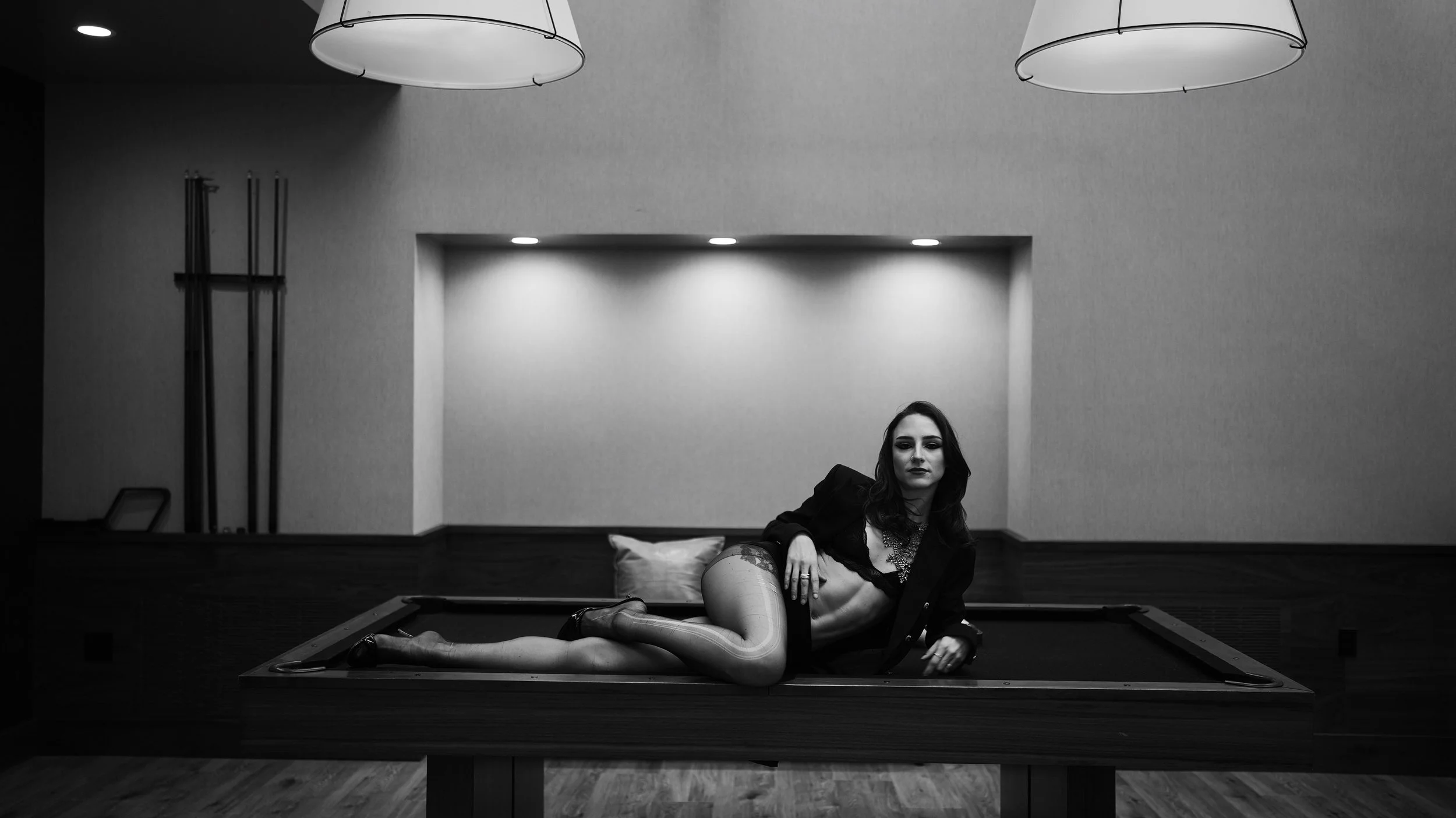 A woman with dark hair, wearing a black jacket and lingerie, lying on a pool table in a dimly lit room with modern ceiling lights.