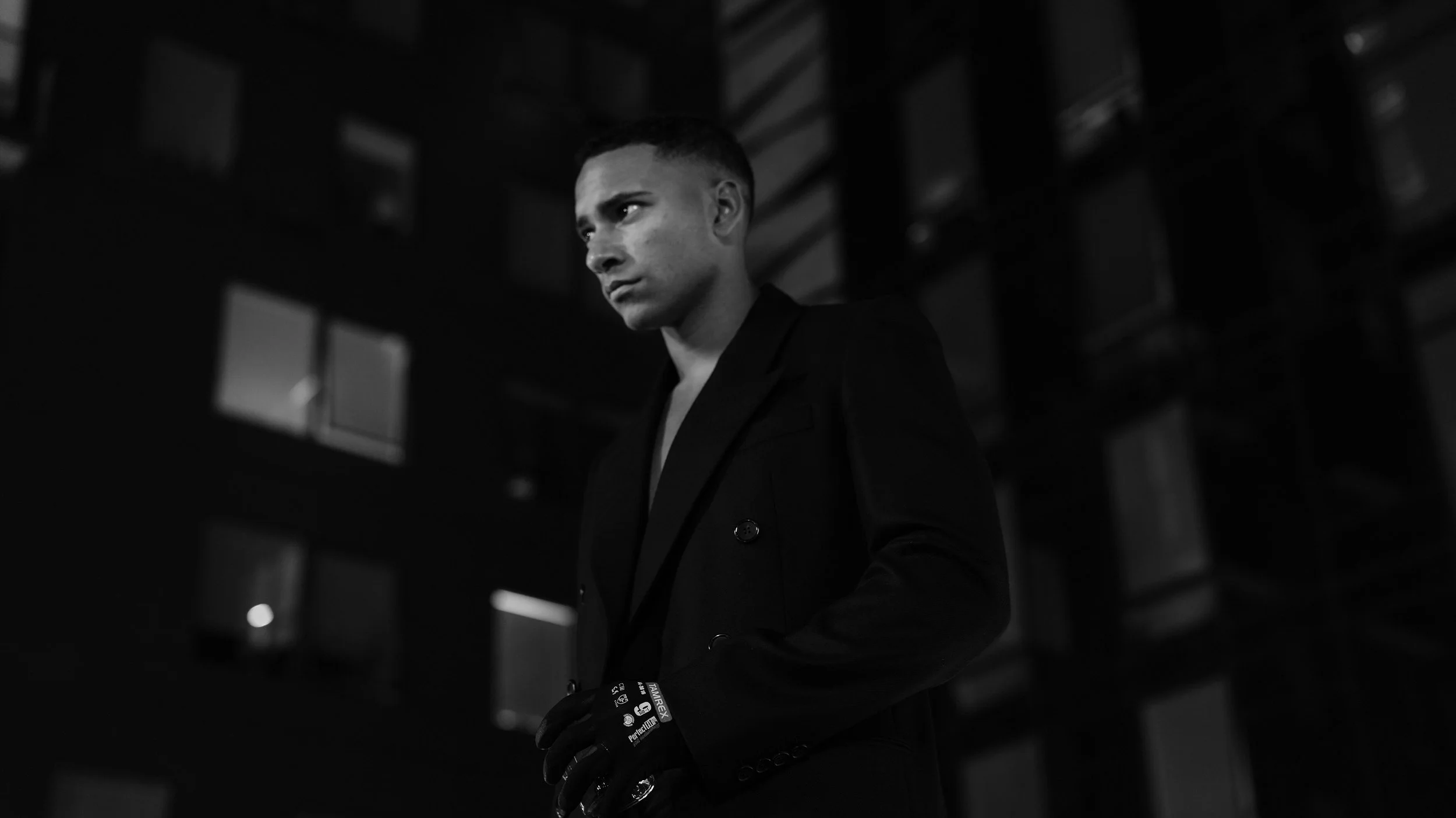 A man dressed in a dark suit and black gloves standing in front of a modern building at night, illuminated by city lights.