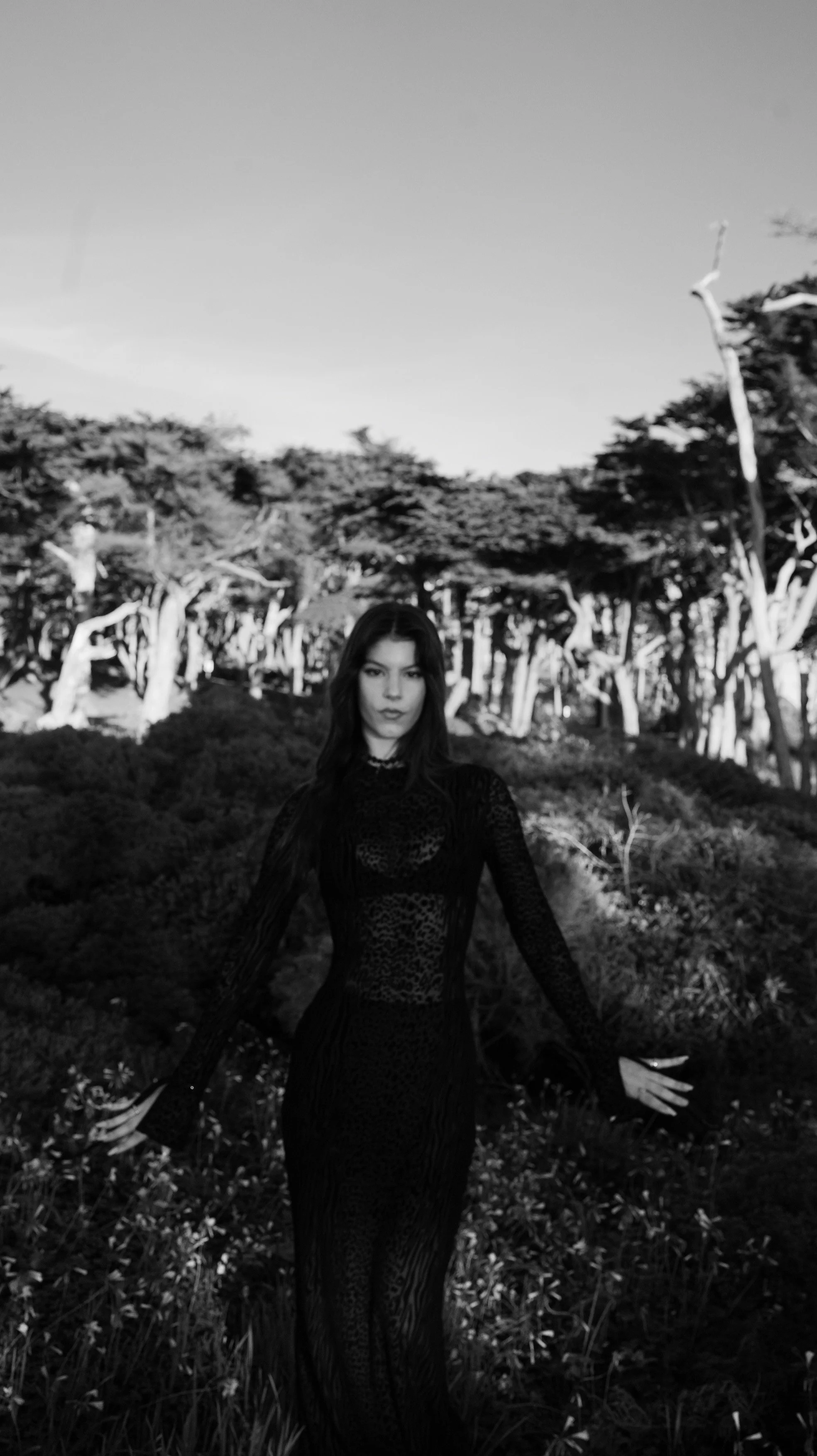 A woman in a long dark dress standing outdoors in a forested area with trees in the background, black and white image.