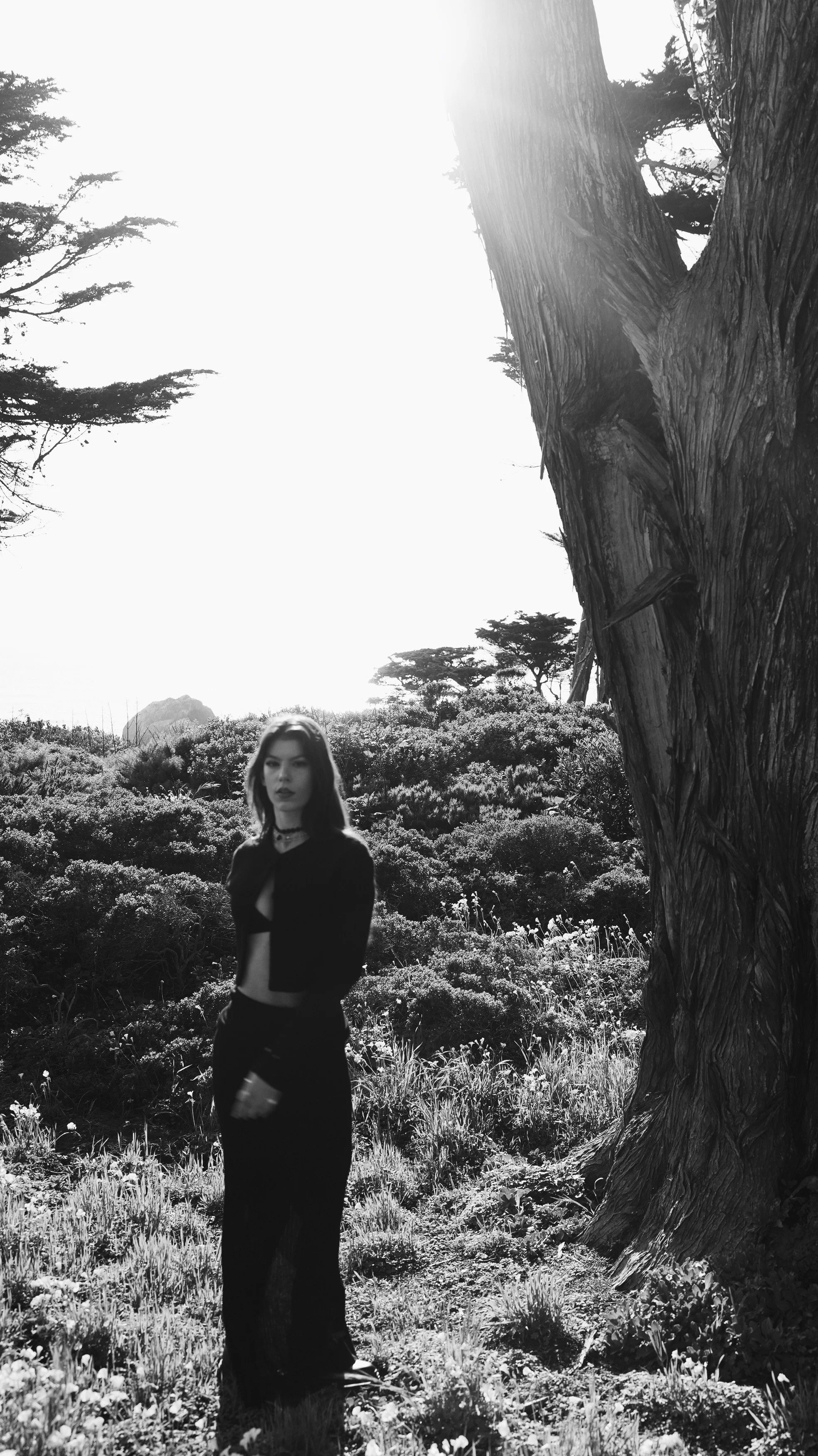 A young woman with shoulder-length hair wearing a black outfit, standing in a natural outdoor setting with trees and bushes, in a black and white photograph.