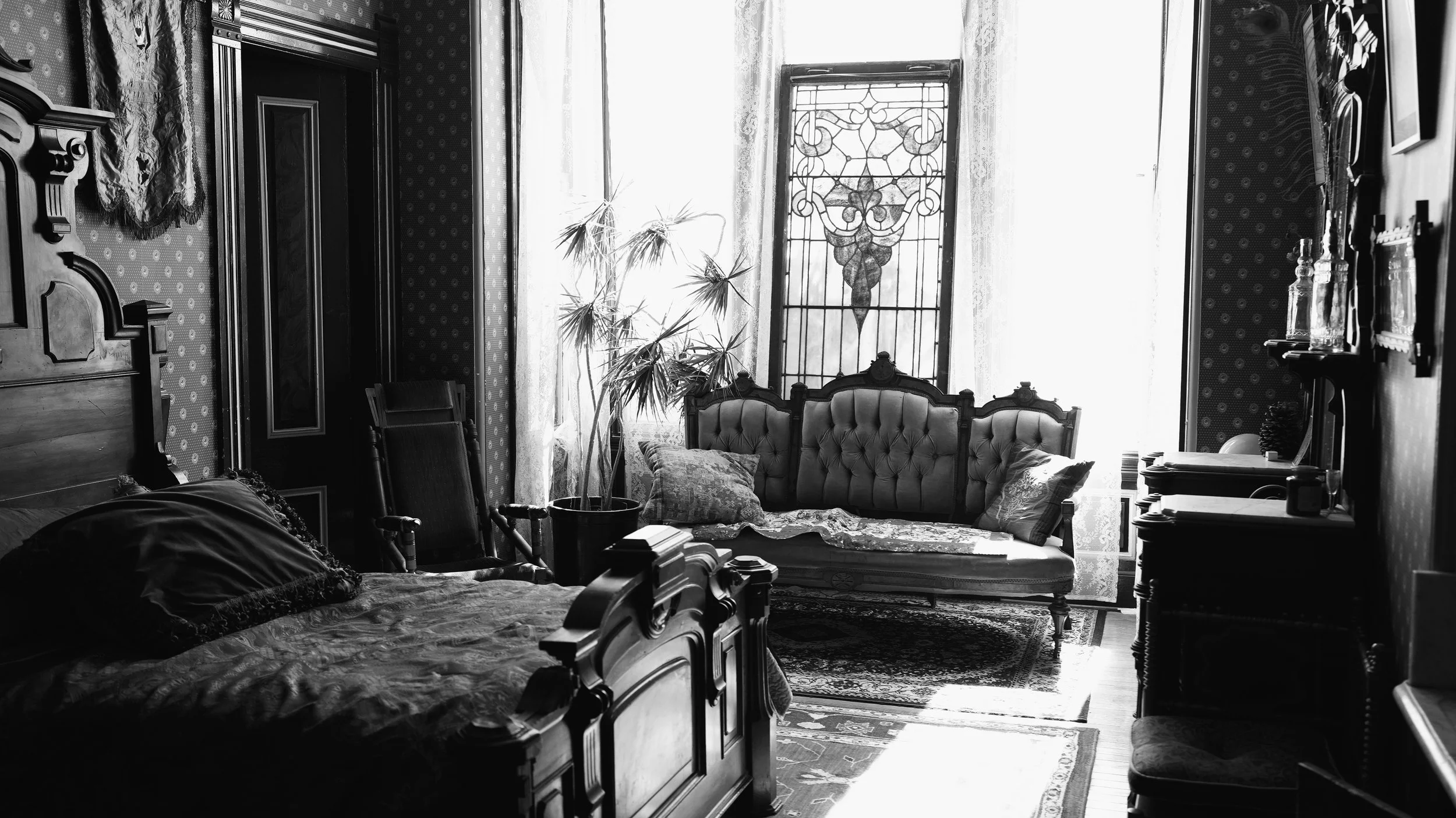 A black and white photo of a vintage living room with a sofa, armchair, and various antique furniture, decorated with patterned wallpaper, lace curtains, and a stained glass window, with a tall plant near the window.