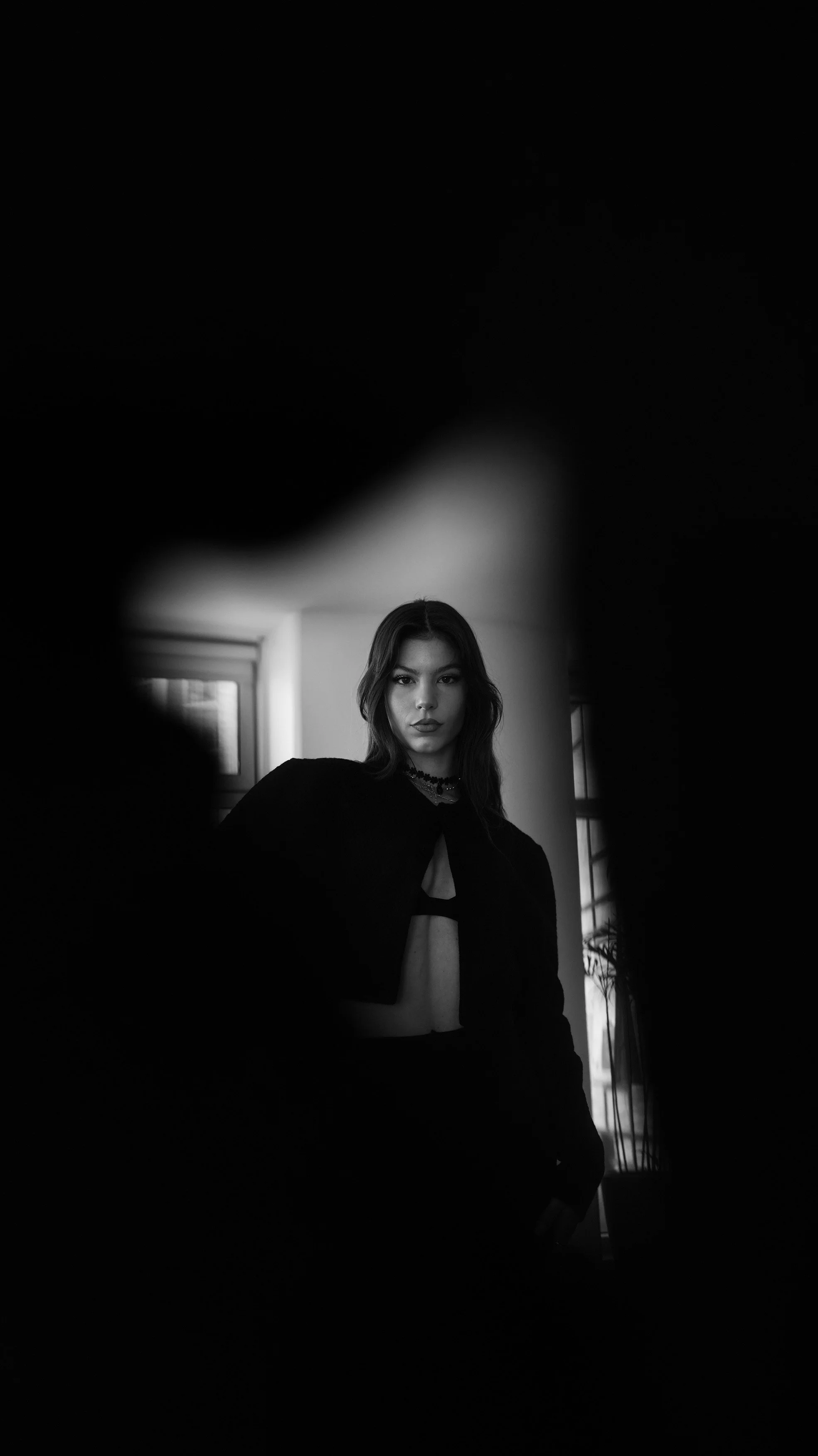 A young woman with dark hair and a serious expression posing indoors, with a window in the background, seen through a dark, irregularly shaped frame.