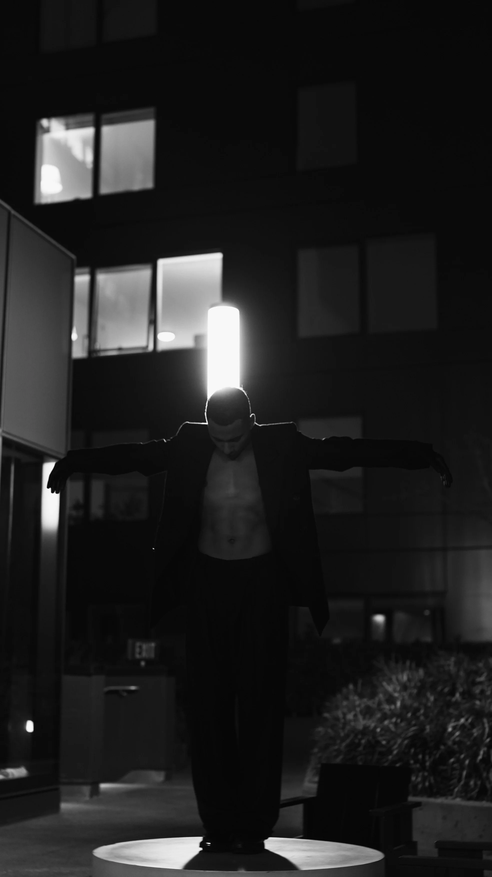 A man with a muscular torso and suit jacket standing on a circular platform with arms outstretched at night, backlit by a vertical light and illuminated windows in a building behind him.