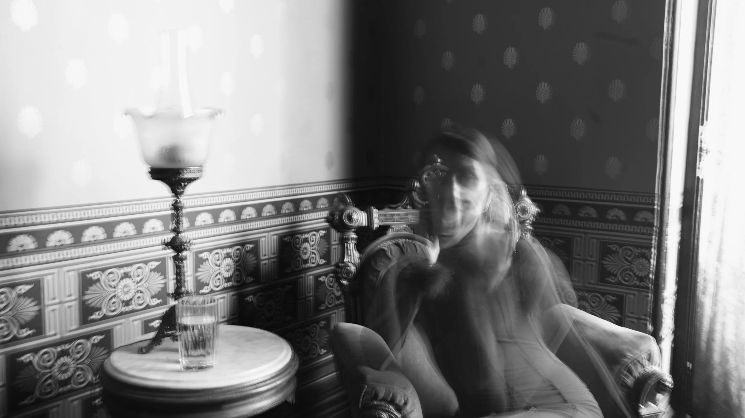 Black and white photo of a person sitting at a table in a vintage-style room with ornate patterned walls, next to a large decorative lamp, with motion blur showing movement or a long exposure shot.