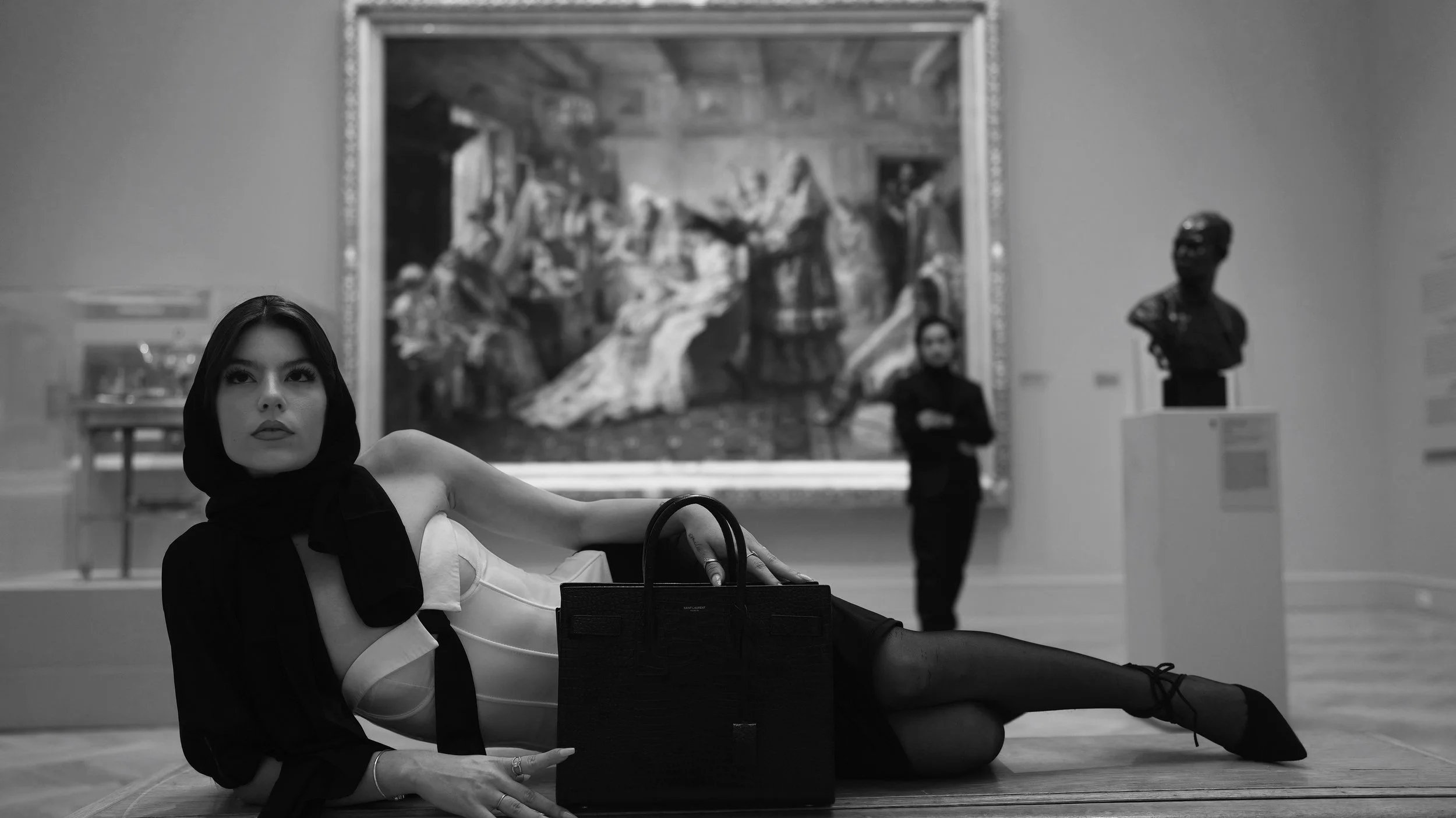 A woman with dark hair and lipstick lying on the floor in an art gallery, wearing a black and white outfit, with a black handbag next to her. Behind her, there is a large painting and a sculpture of a bust. A man in a dark suit with crossed arms is i