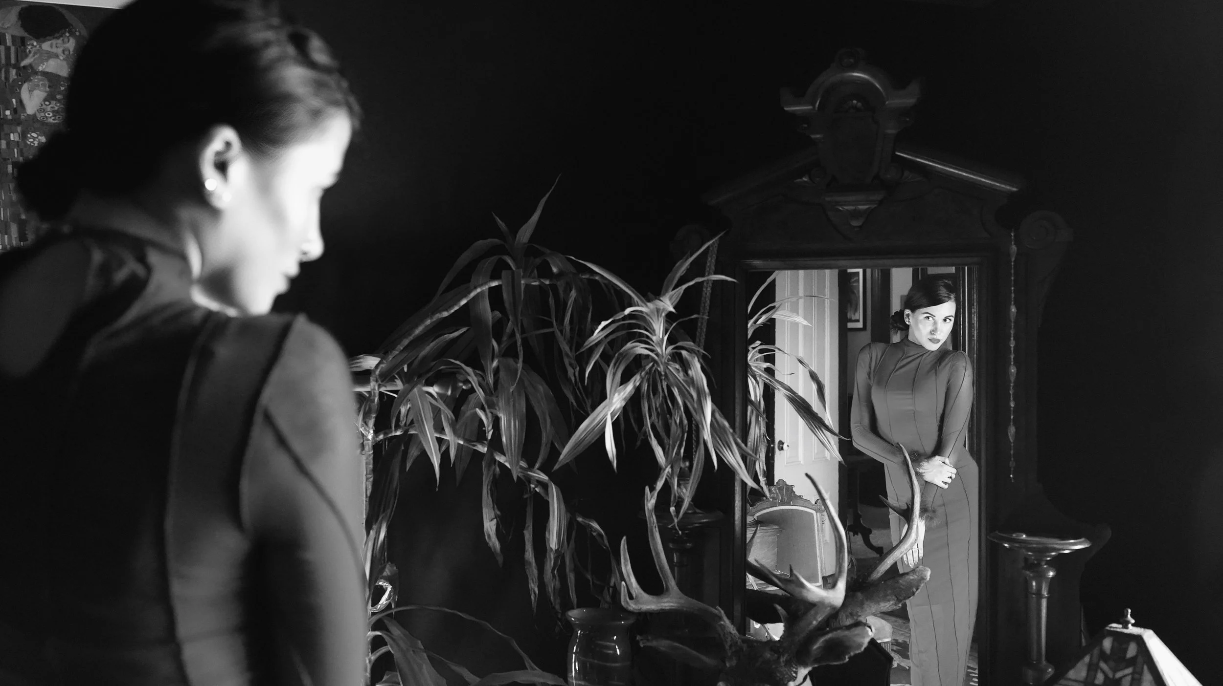 A woman in a dress looking at her reflection in a mirror in a room with plants and furniture, seen in a reflection.