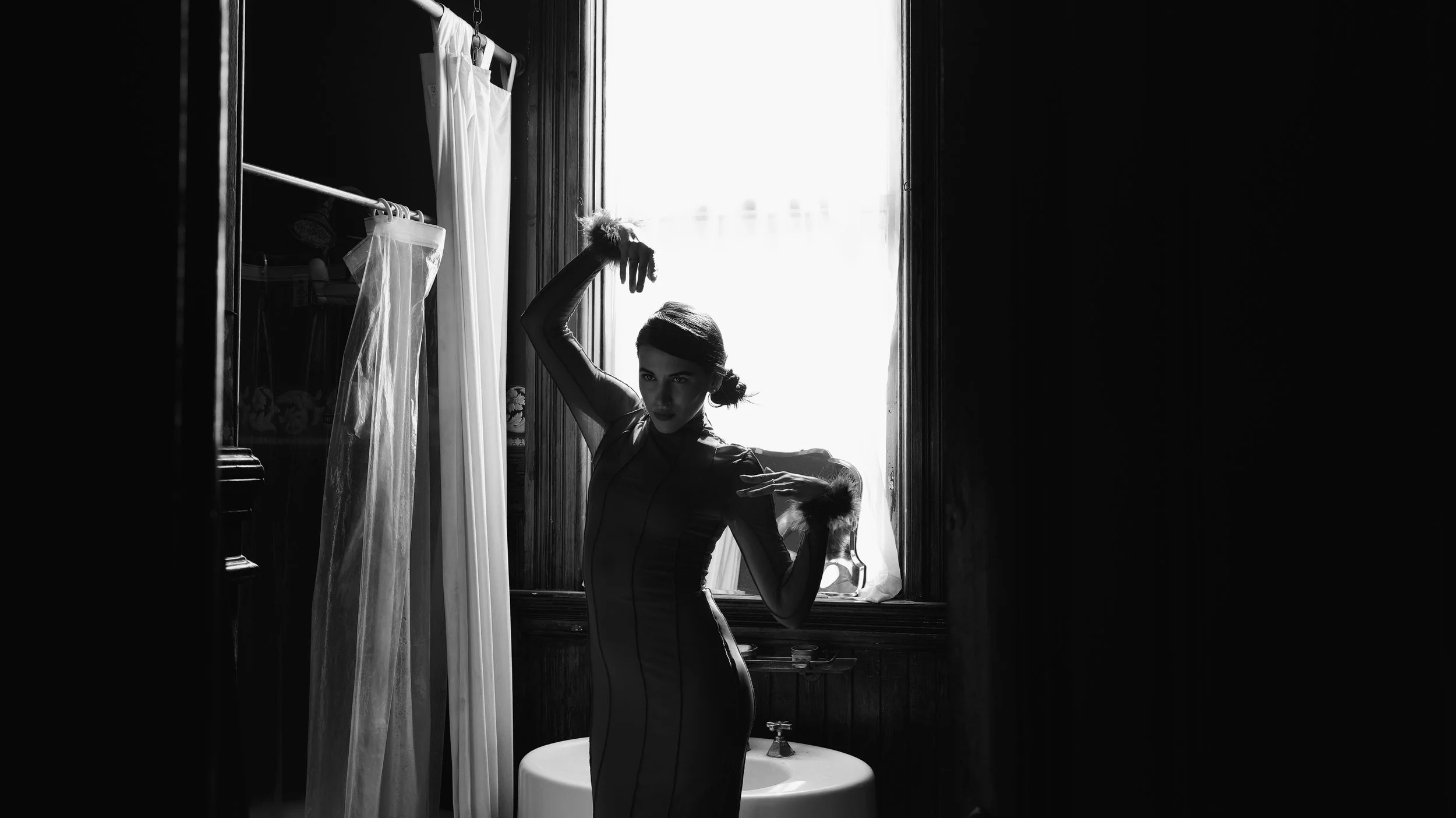 A woman in vintage-style clothing standing in front of a window with bright light coming through, black and white photo.