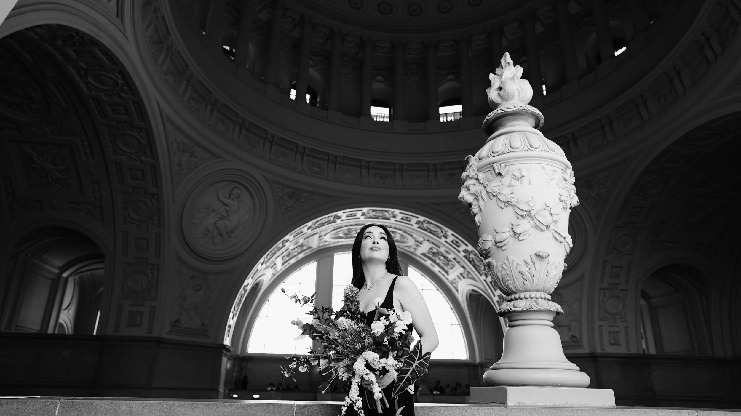 A woman in a black dress holding a bouquet of flowers inside a grand, ornate building with high ceilings and decorative architecture, including large windows and sculptures.