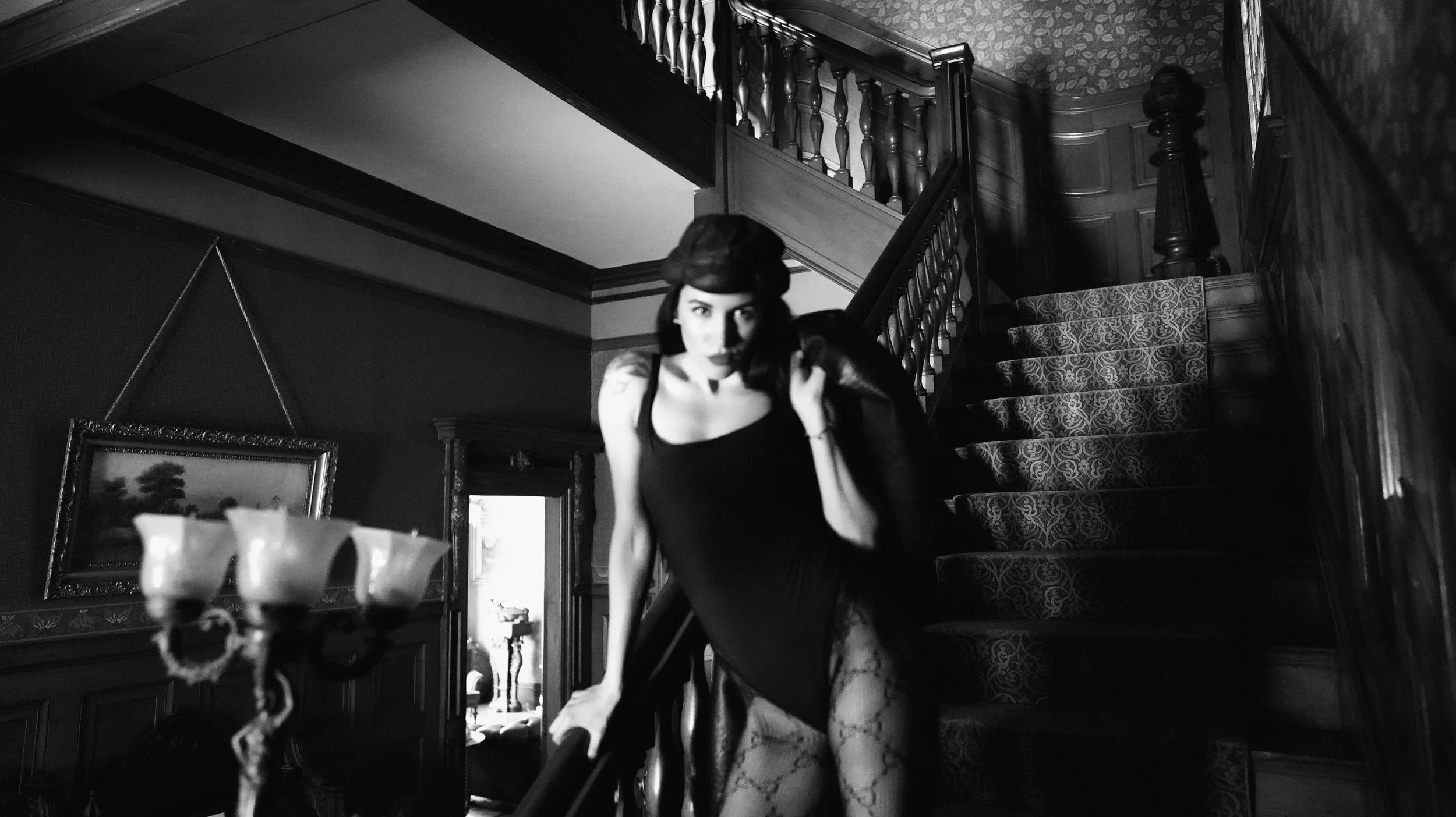A black and white photo of a woman with dark hair wearing a black dress, posing on a staircase in a vintage styled room with wood paneling and ornate furniture.