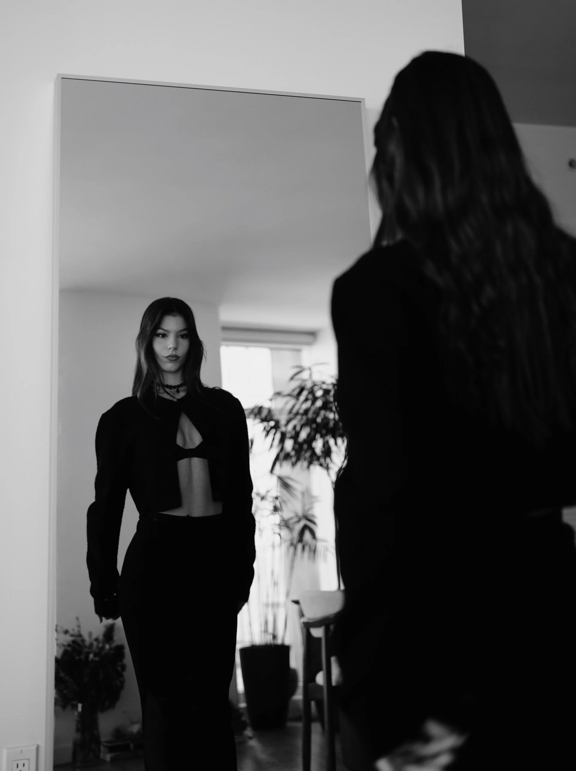A woman with long hair looking at the camera through a mirror, wearing dark clothing with a cutout top, in a room with houseplants and a chair.