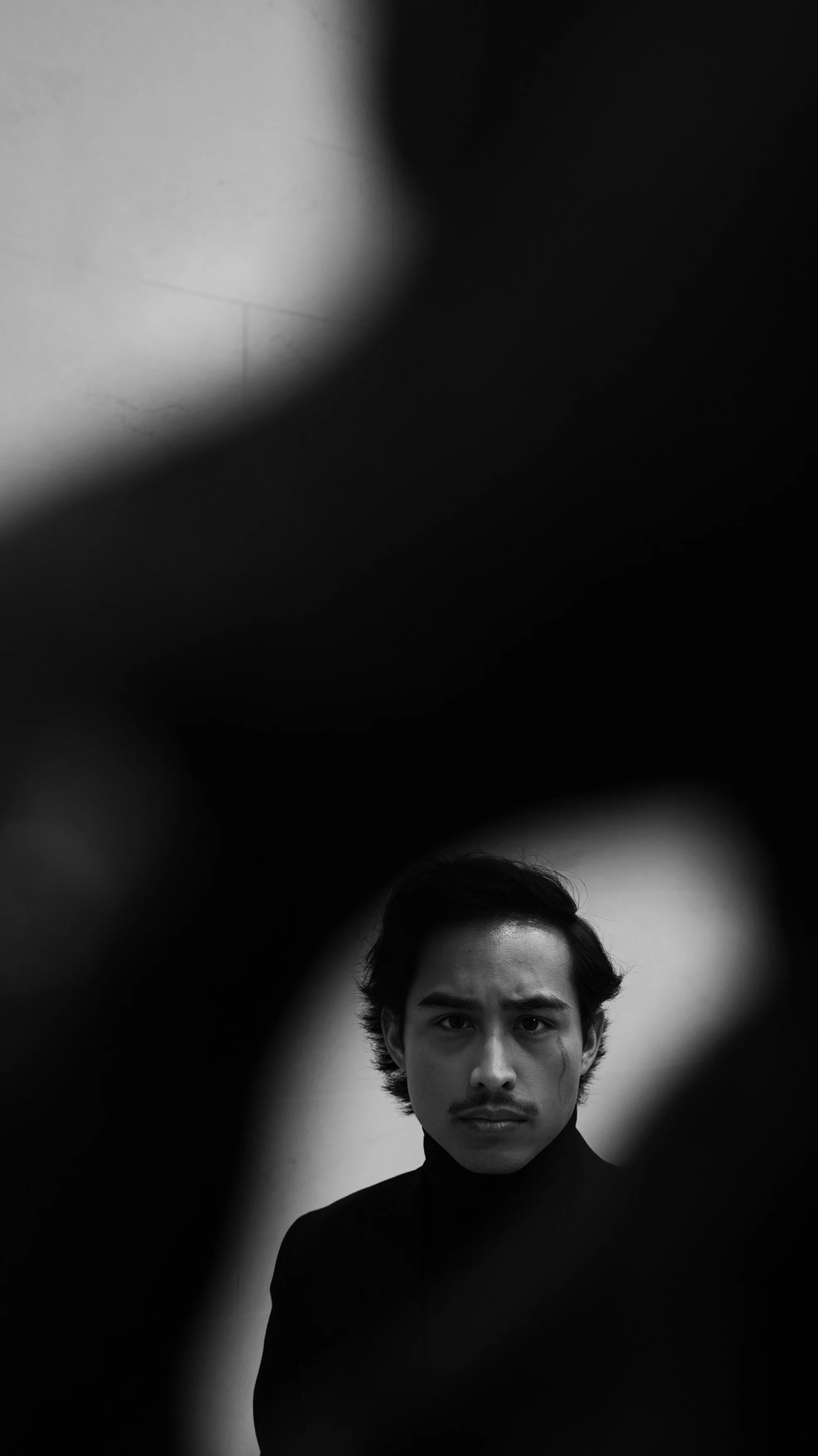 Black and white photo of a young man with dark hair, wearing a black shirt, staring into the camera, with a blurred background and a light source creating a halo effect around his head.