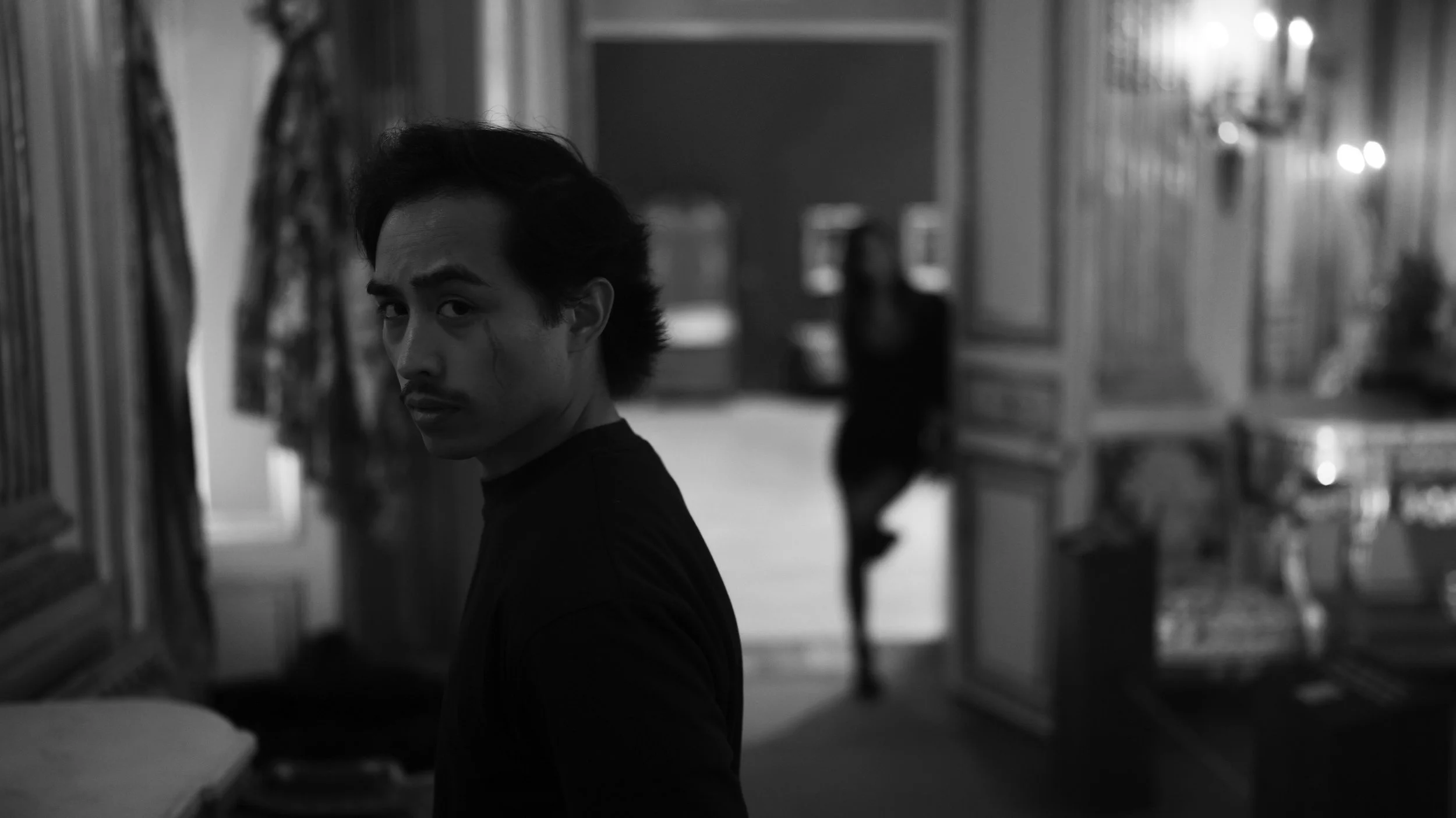 A black-and-white photo of a young man with dark hair and a mustache, looking at the camera, in a store aisle with shelves and blurred background.