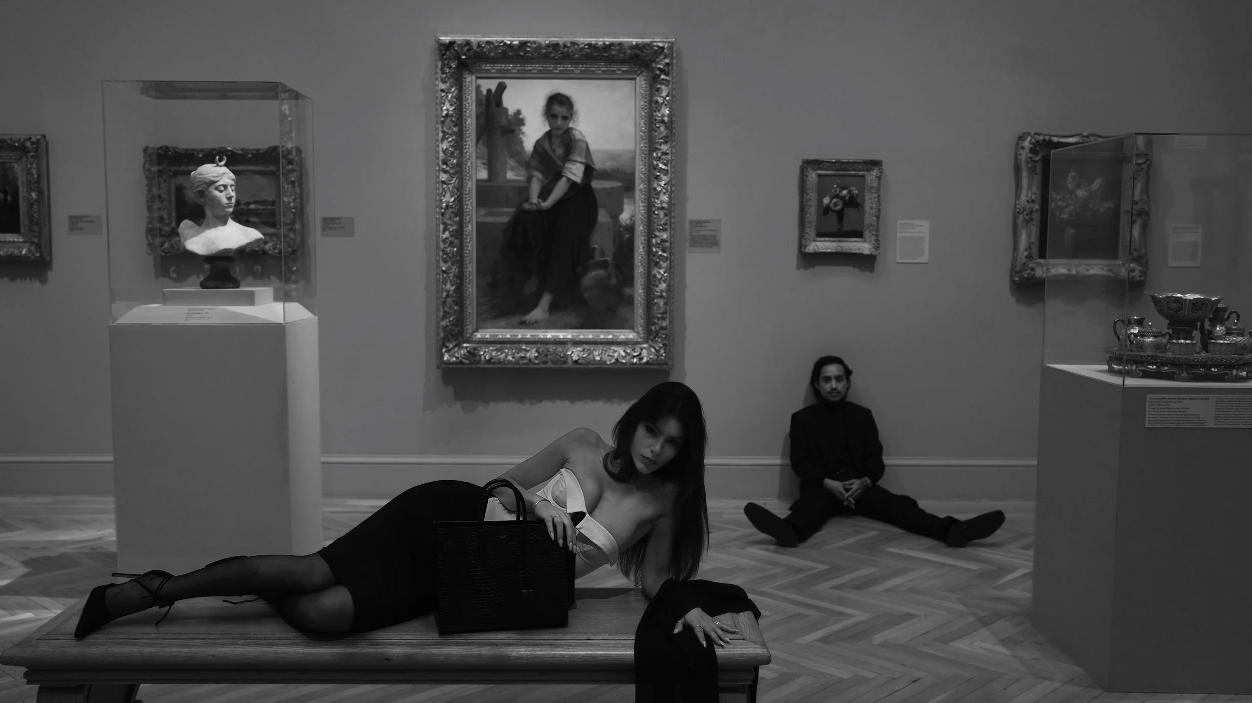 Two women are posing on the floor in front of a gallery wall with paintings and a bust sculpture, one woman lying on a bench and the other sitting on the floor. Two men are sitting against the wall in the background.