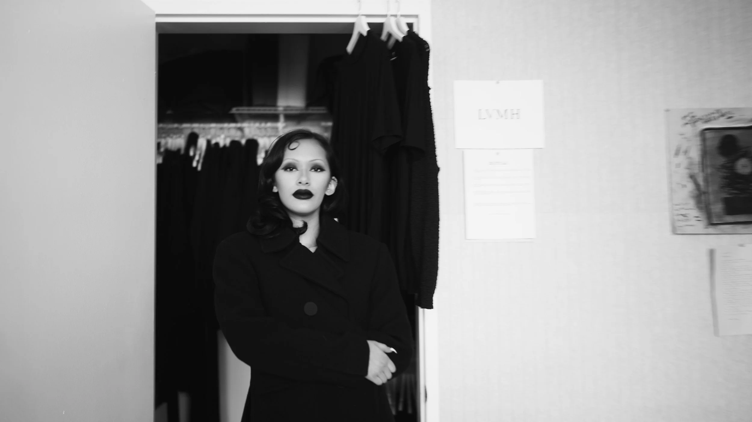Black-and-white photo of a woman with dark wavy hair, wearing dark lipstick and a dark coat, standing in front of a doorway in an indoor setting.