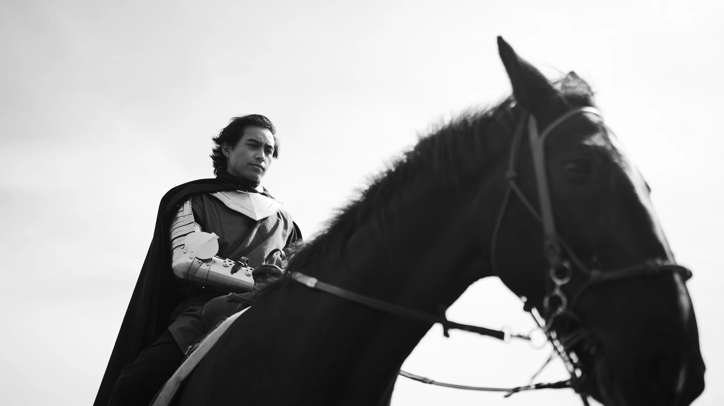 Black and white photo of a man in armor and a cape riding a horse, looking contemplative.
