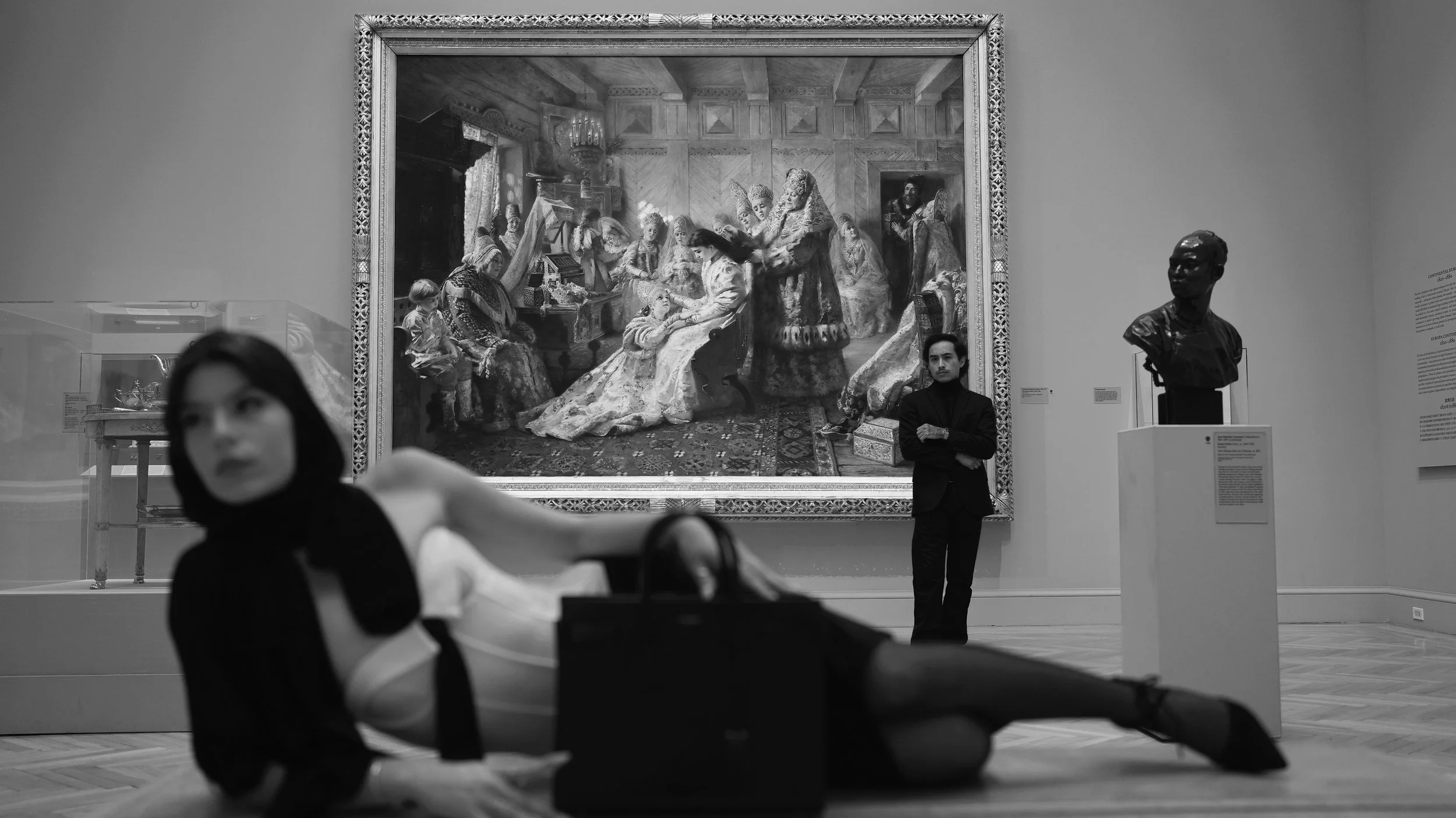Black and white photo of an art gallery showing a large framed painting of a historical scene, a bust sculpture on a pedestal, and two museum visitors observing the exhibits. The foreground has an out-of-focus mannequin or figure of a person lying do