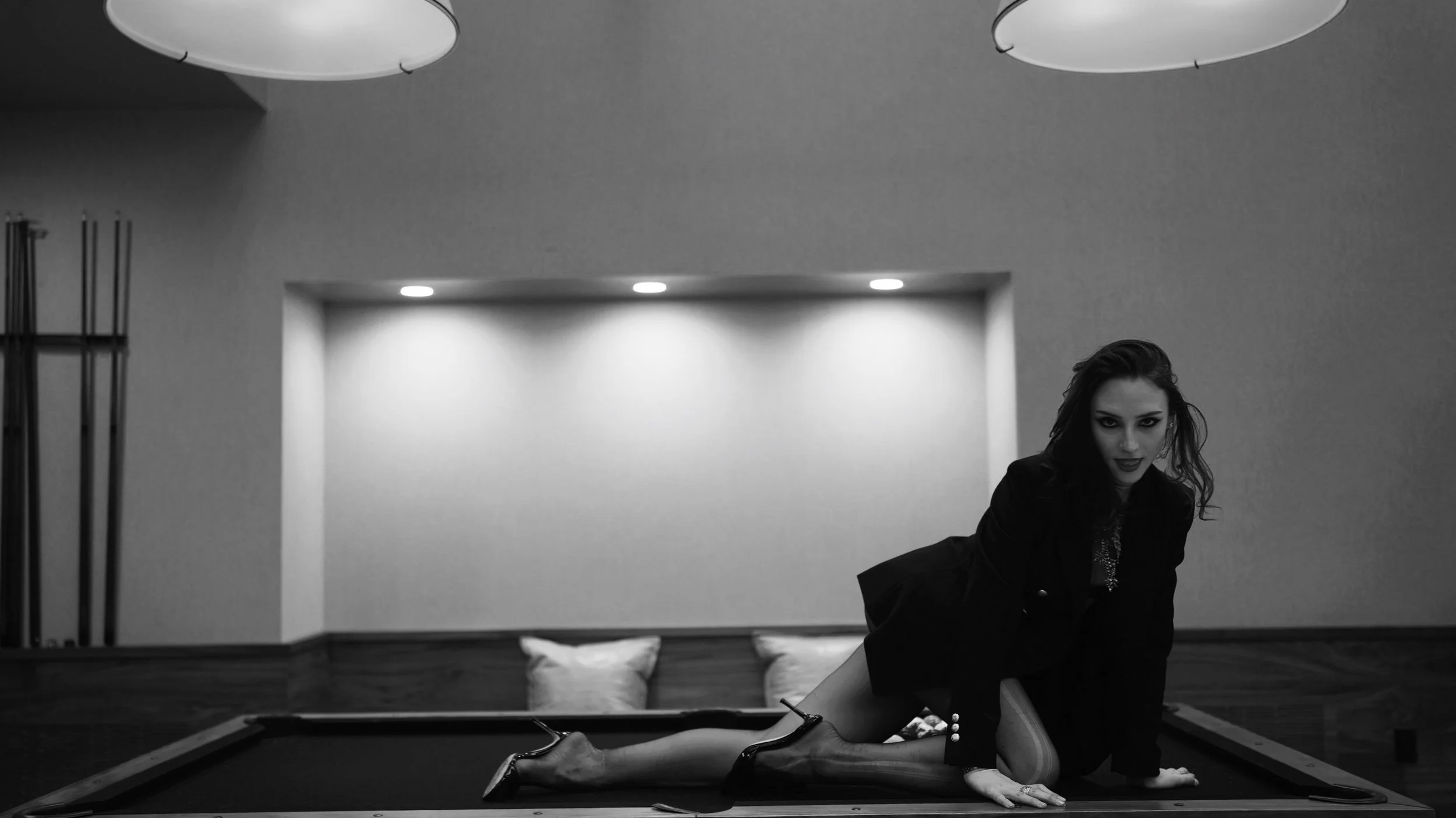 A woman with dark hair and makeup leaning on a pool table in a modern room with minimalist decor, including a rectangular alcove with ceiling lights and hanging lamps.