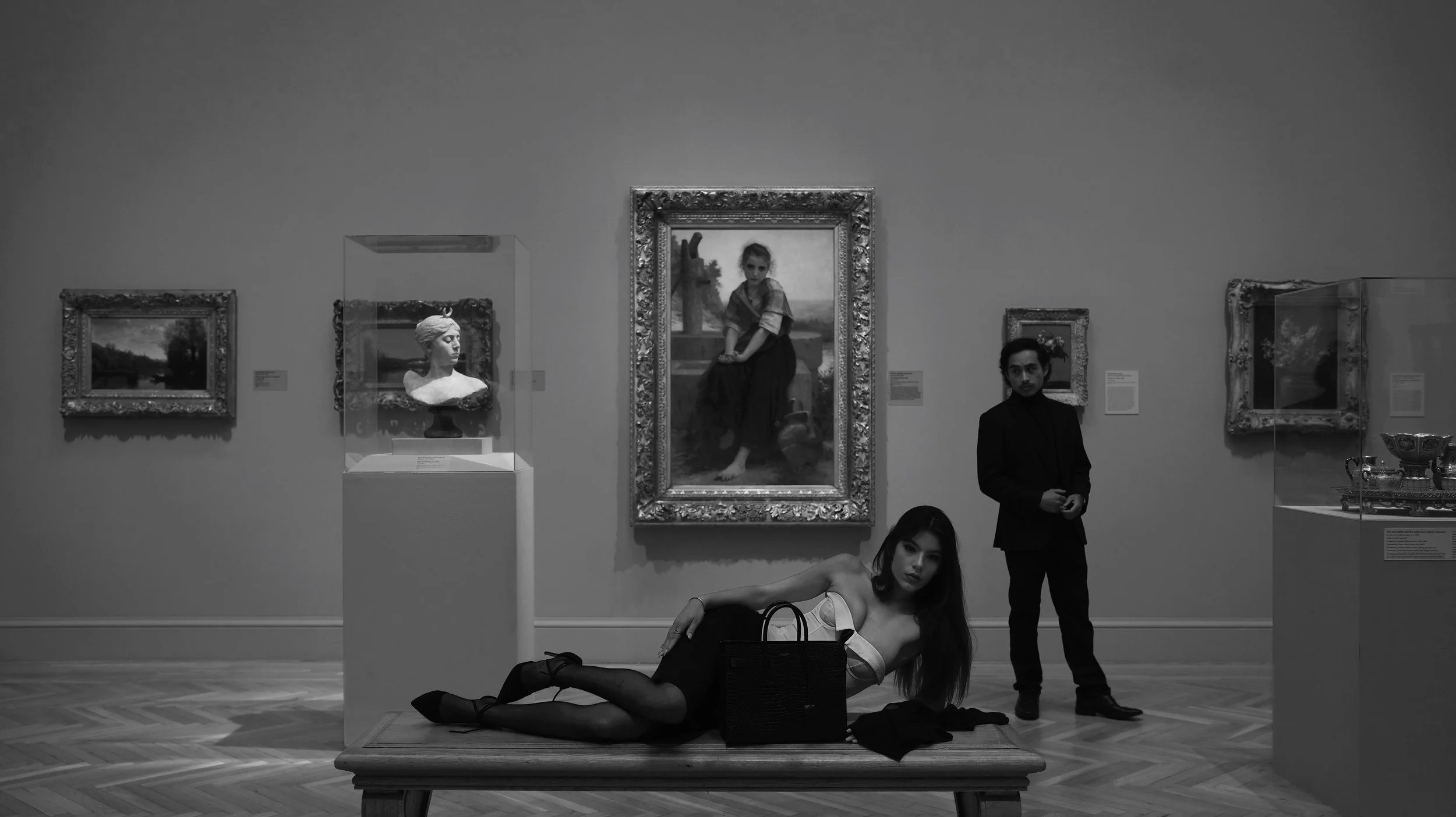 Two young women with long hair, dressed in fashionable outfits, pose on the floor of an art gallery while two men stand nearby. The gallery wall displays several framed paintings and sculptures, including busts and a portrait of a girl, all with orna