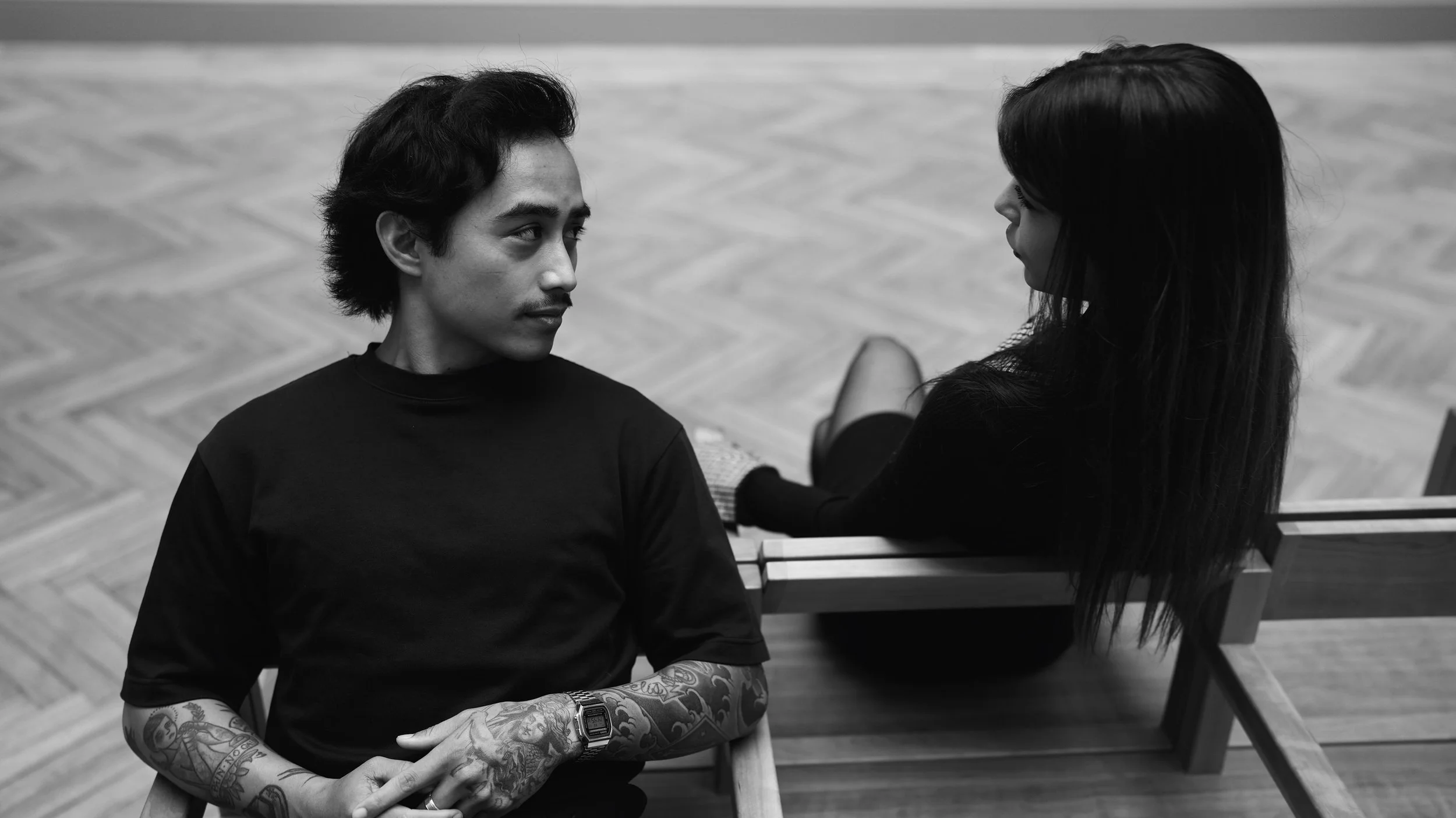 A young man with tattoos on his arms and a wristwatch is sitting on a wooden bench, looking at a young woman with long dark hair sitting on a similar bench nearby, in a room with a herringbone wooden floor.