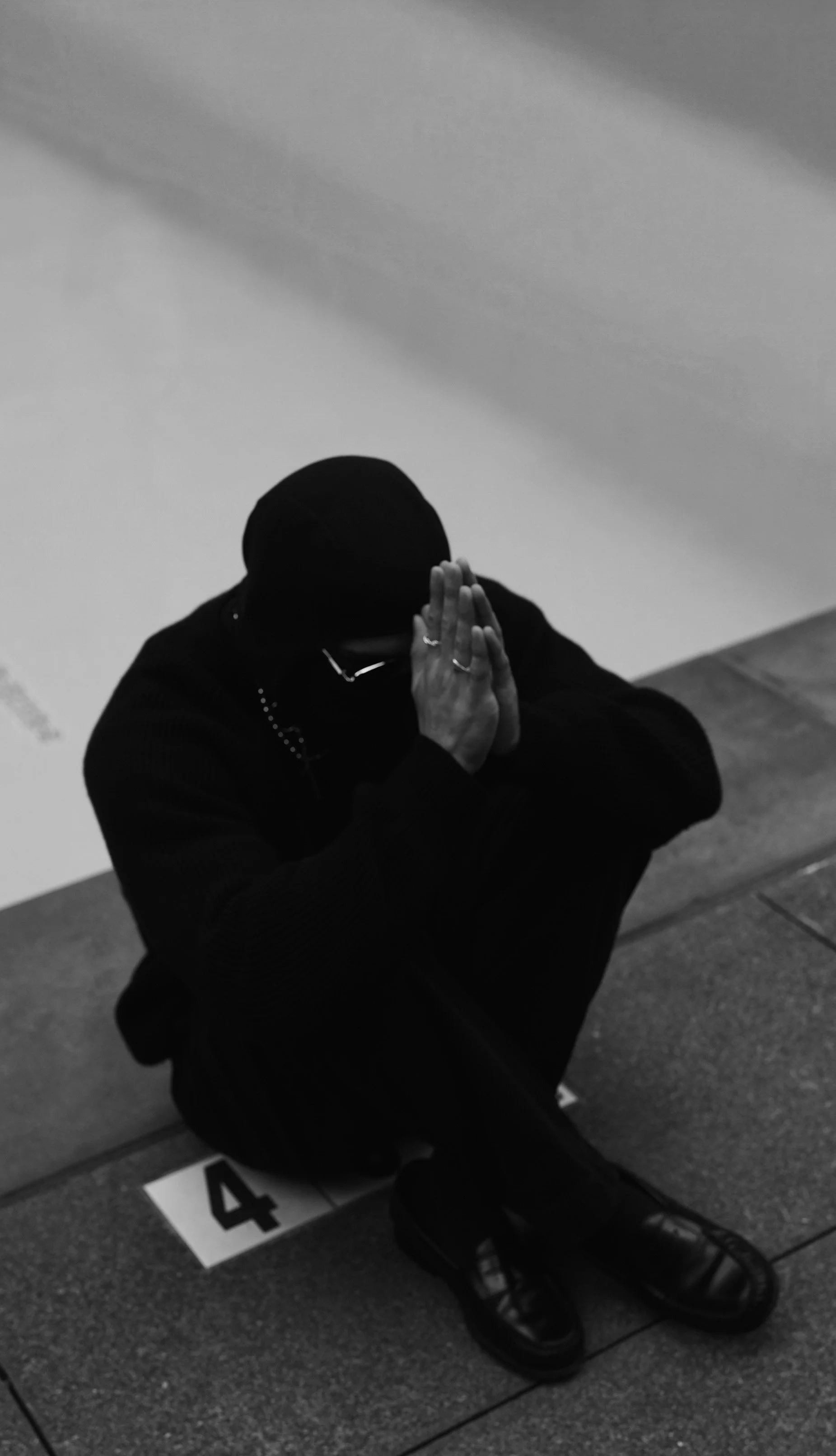 Black-and-white photo of a person kneeling on a sidewalk, wearing dark clothing, with hands pressed together in prayer, head bowed, and face obscured by a hood or hat. The person is sitting on a ground marked with the number 4.