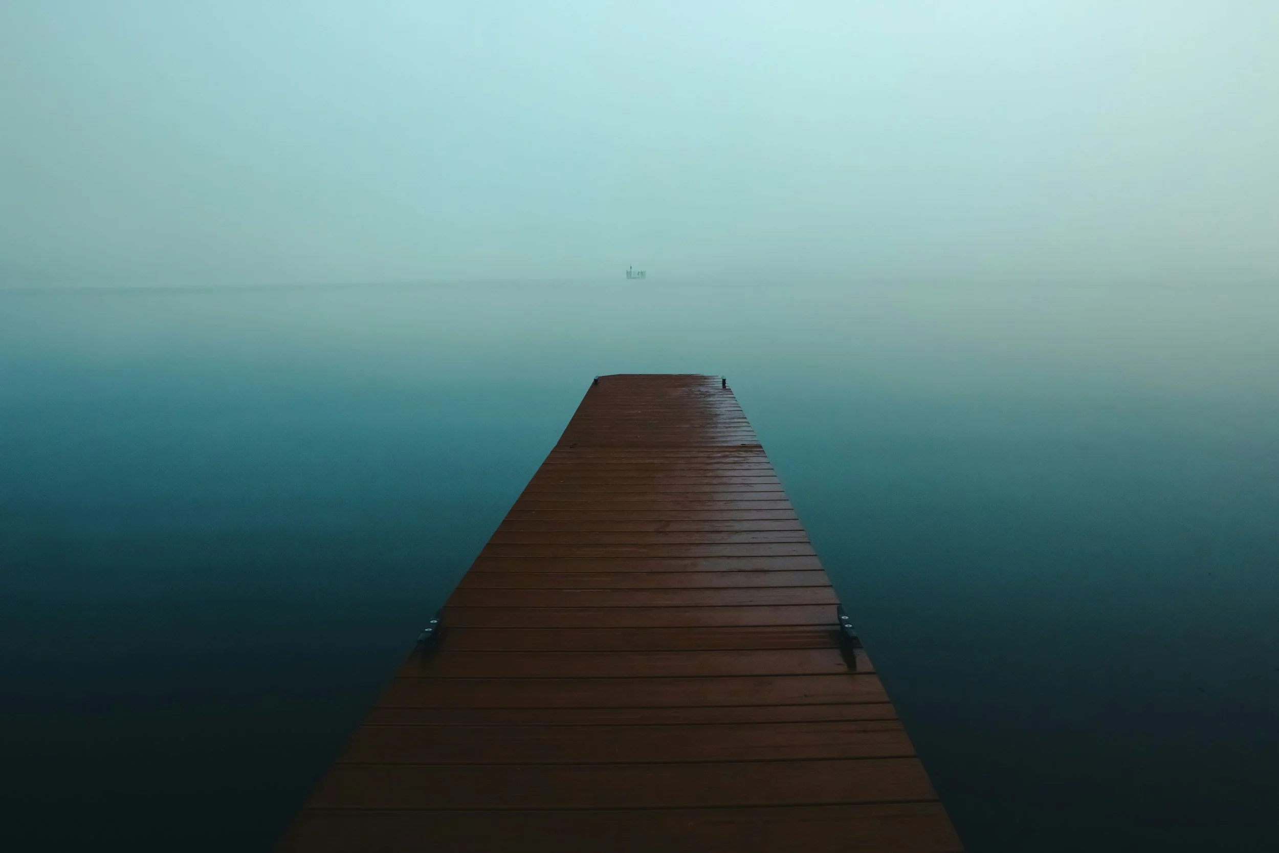 A wooden dock extending into a calm, foggy body of water with a distant boat on the horizon.