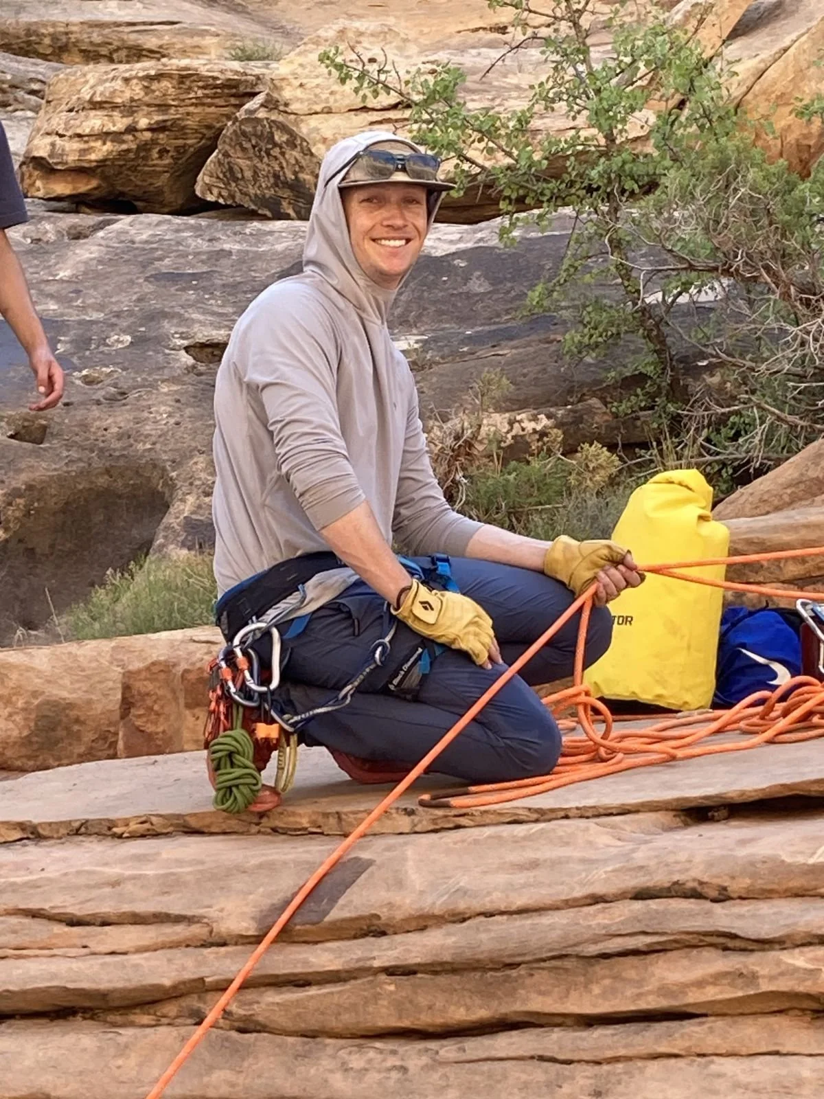 A man in outdoor gear, wearing a hat and gloves, is kneeling on a rocky surface in a desert landscape with large rocks and sparse vegetation, holding a climbing rope.