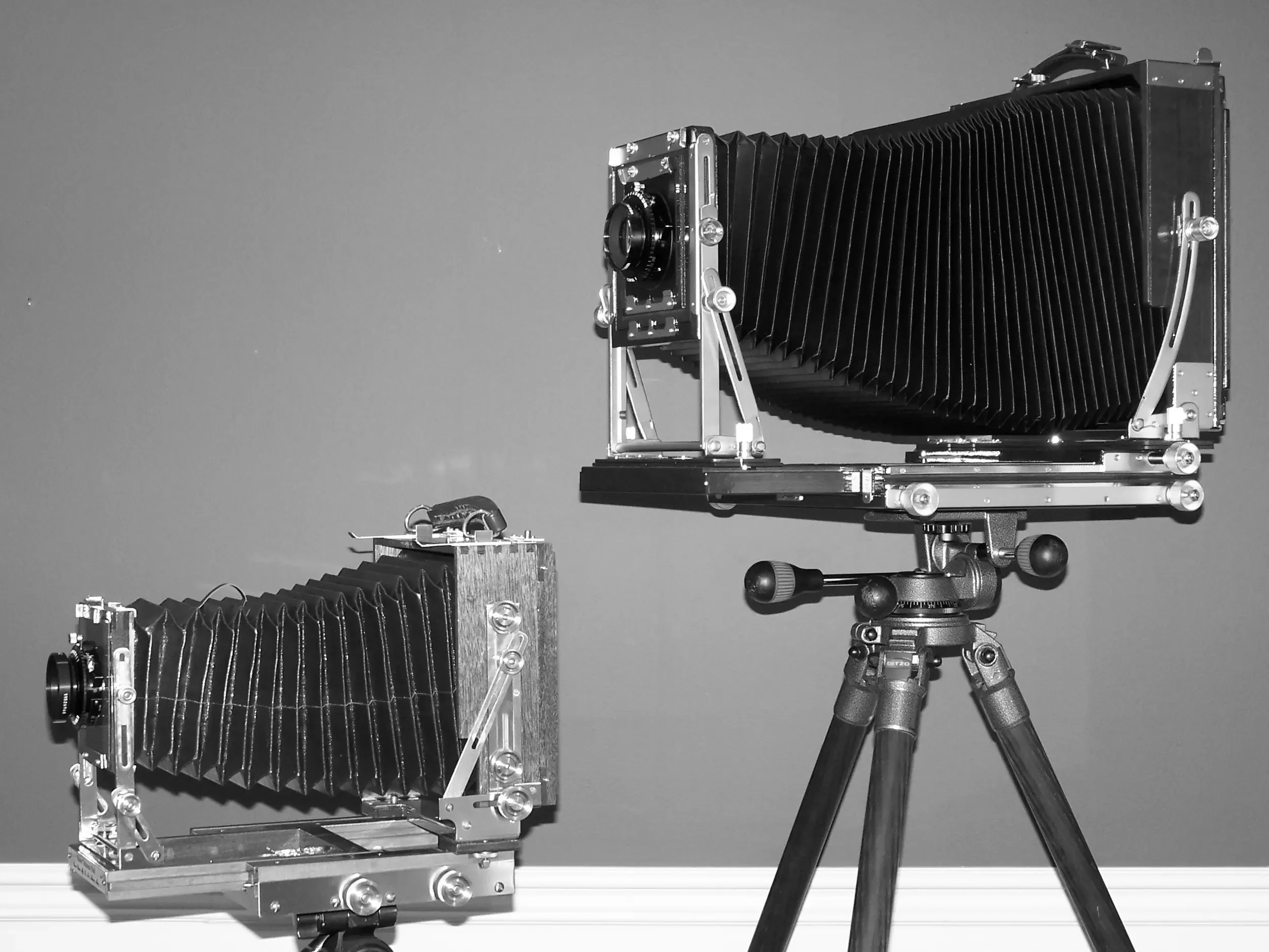 Black and white photo of large-format camera equipment, including a camera with a bellows and a tripod, set up indoors against a plain wall.