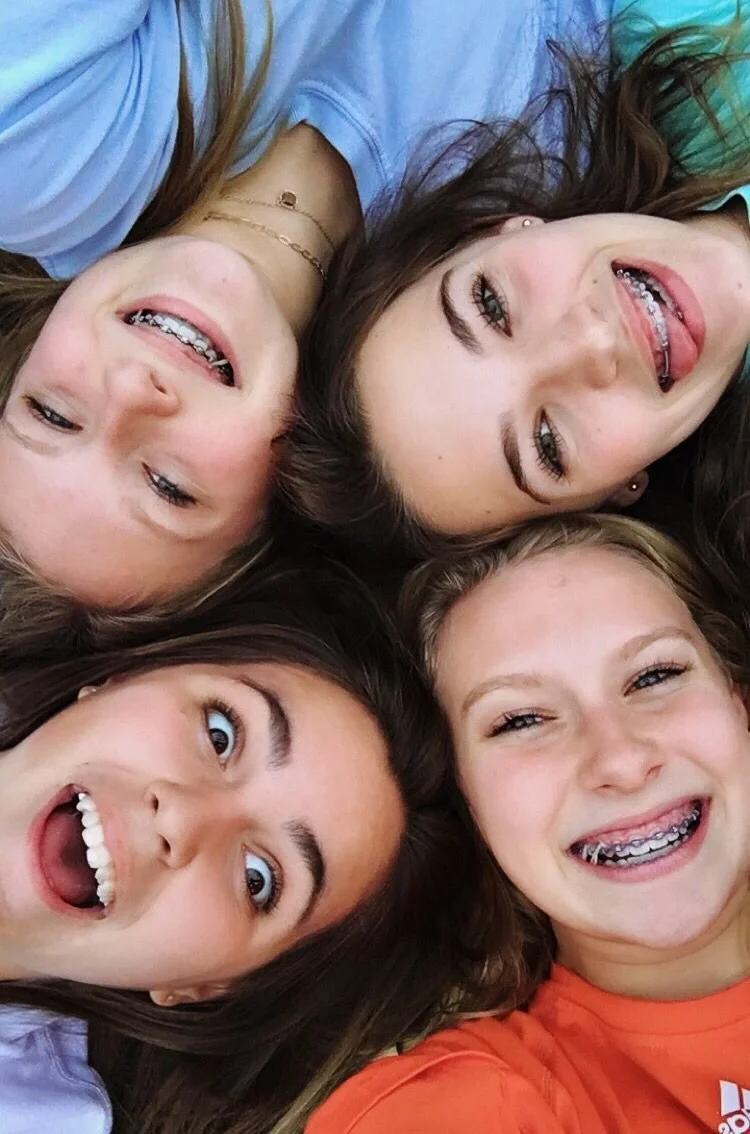 Four young people lying in a circle taking a selfie, all smiling or making funny faces, with braces visible on their teeth.