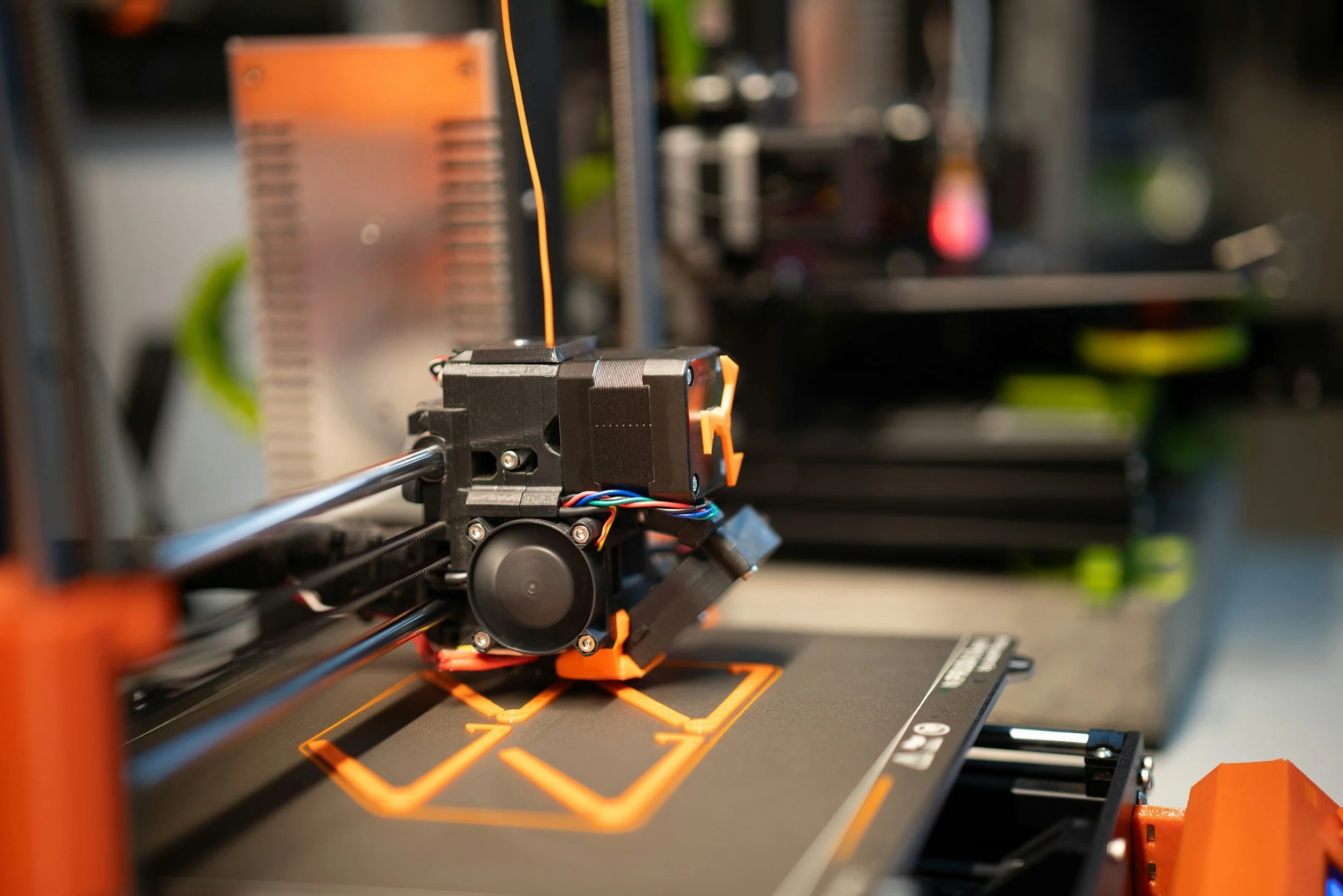 Close-up of a 3D printer extruder printing orange filament on a black build plate with orange outlines.