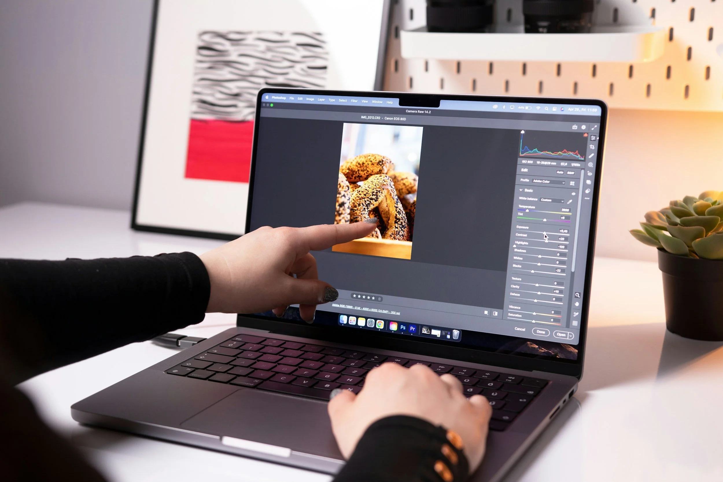 A person is editing a photo of baked goods on a laptop screen using photo editing software in a workspace. The workspace has a patterned poster and a small potted succulent plant.