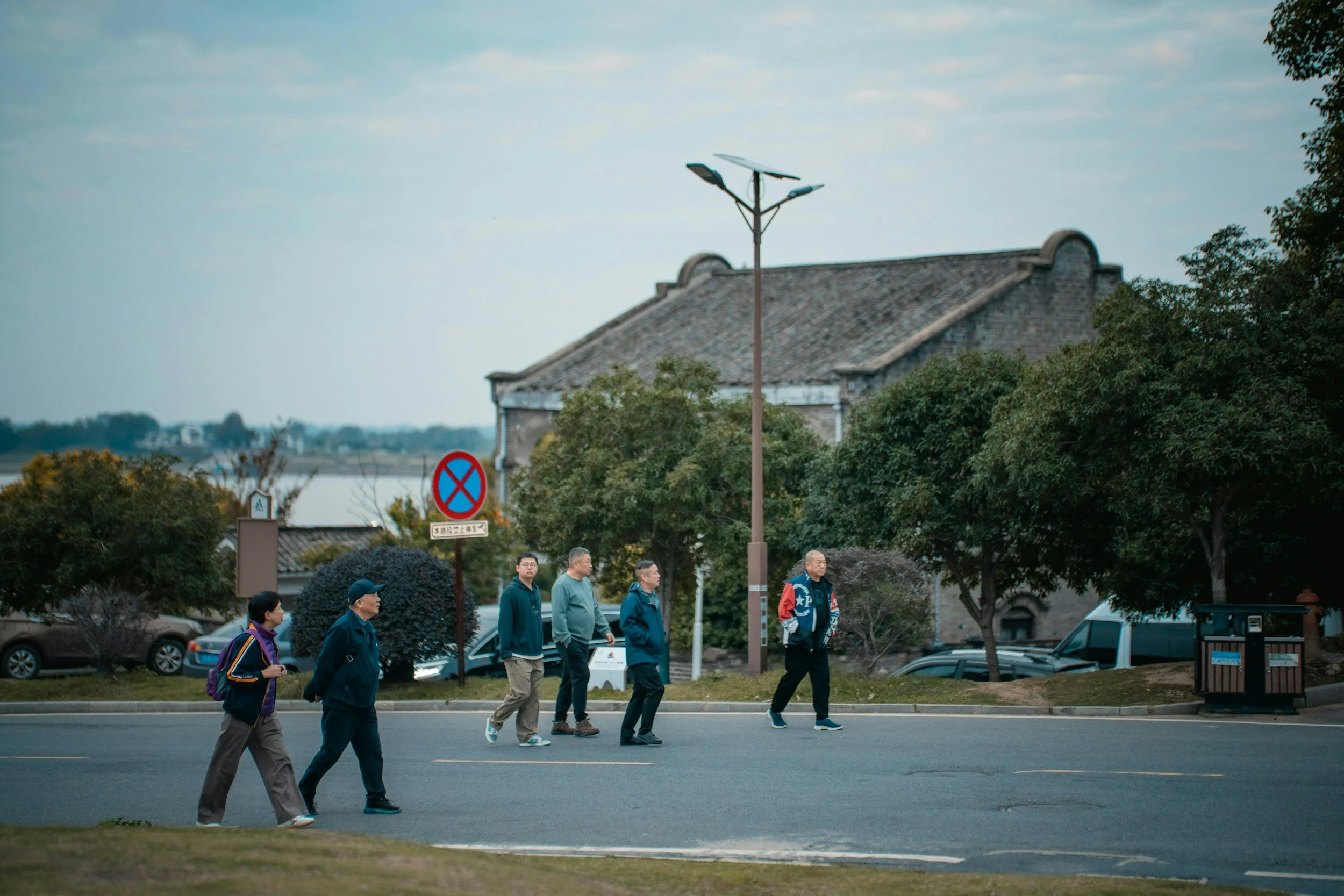 A group of people walking on a city street near a crosswalk, with trees, parked cars, and a building with a tiled roof in the background.