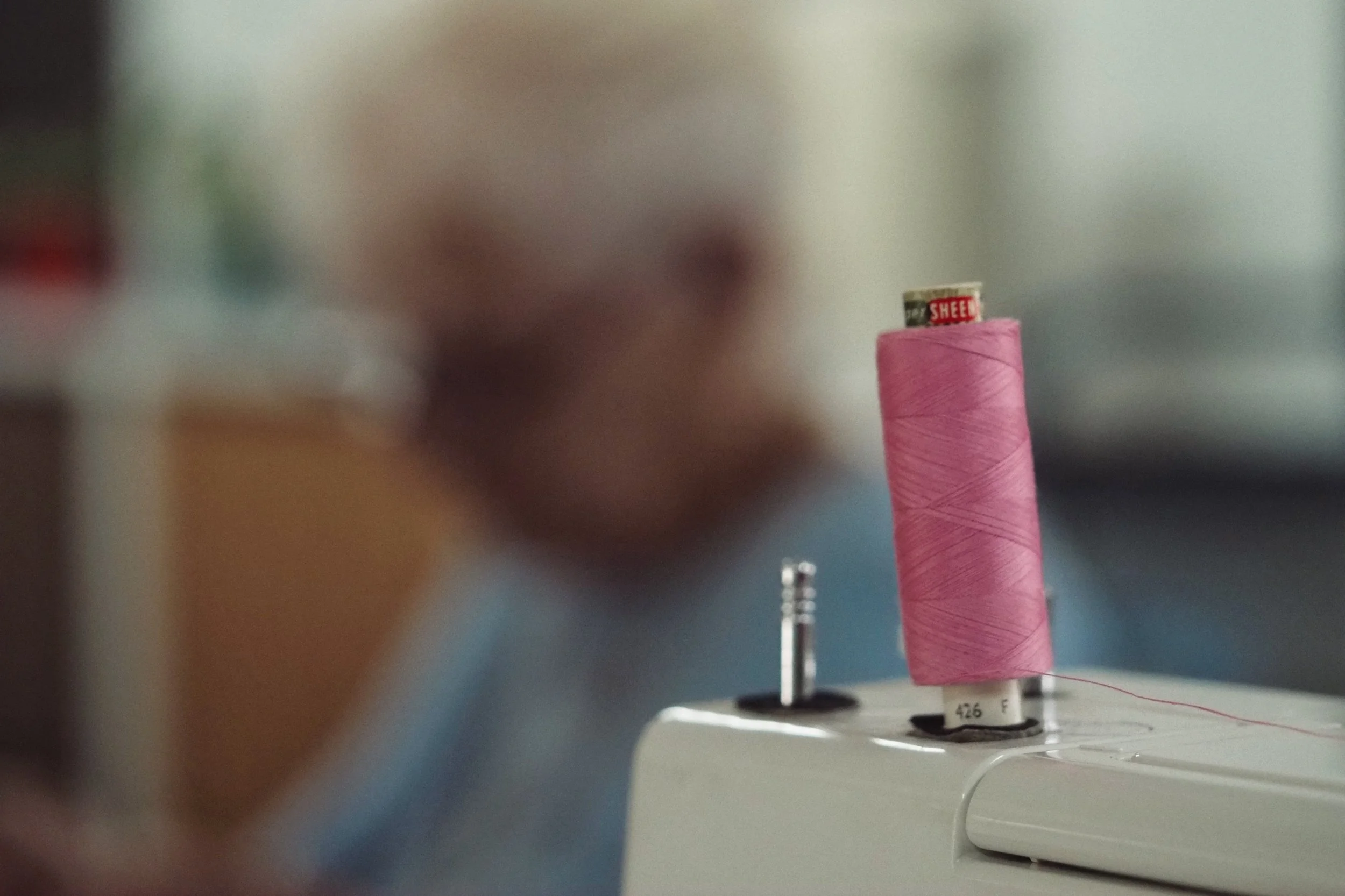 Close-up of a pink spool of thread on a sewing machine with a sewing needle, with a blurred person in the background.