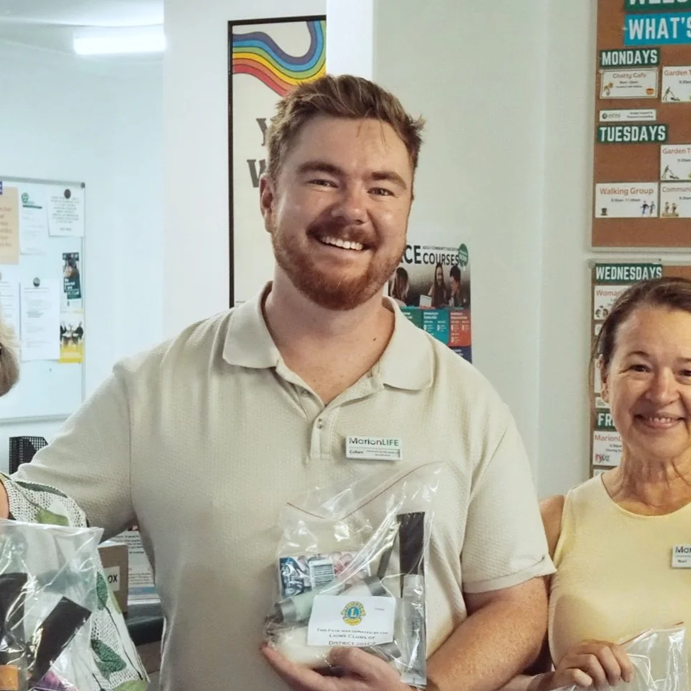 A smiling man with a beard and glasses holding a small gift bag, standing next to a woman in a light yellow shirt, both indoors with posters and notices on the wall behind them.